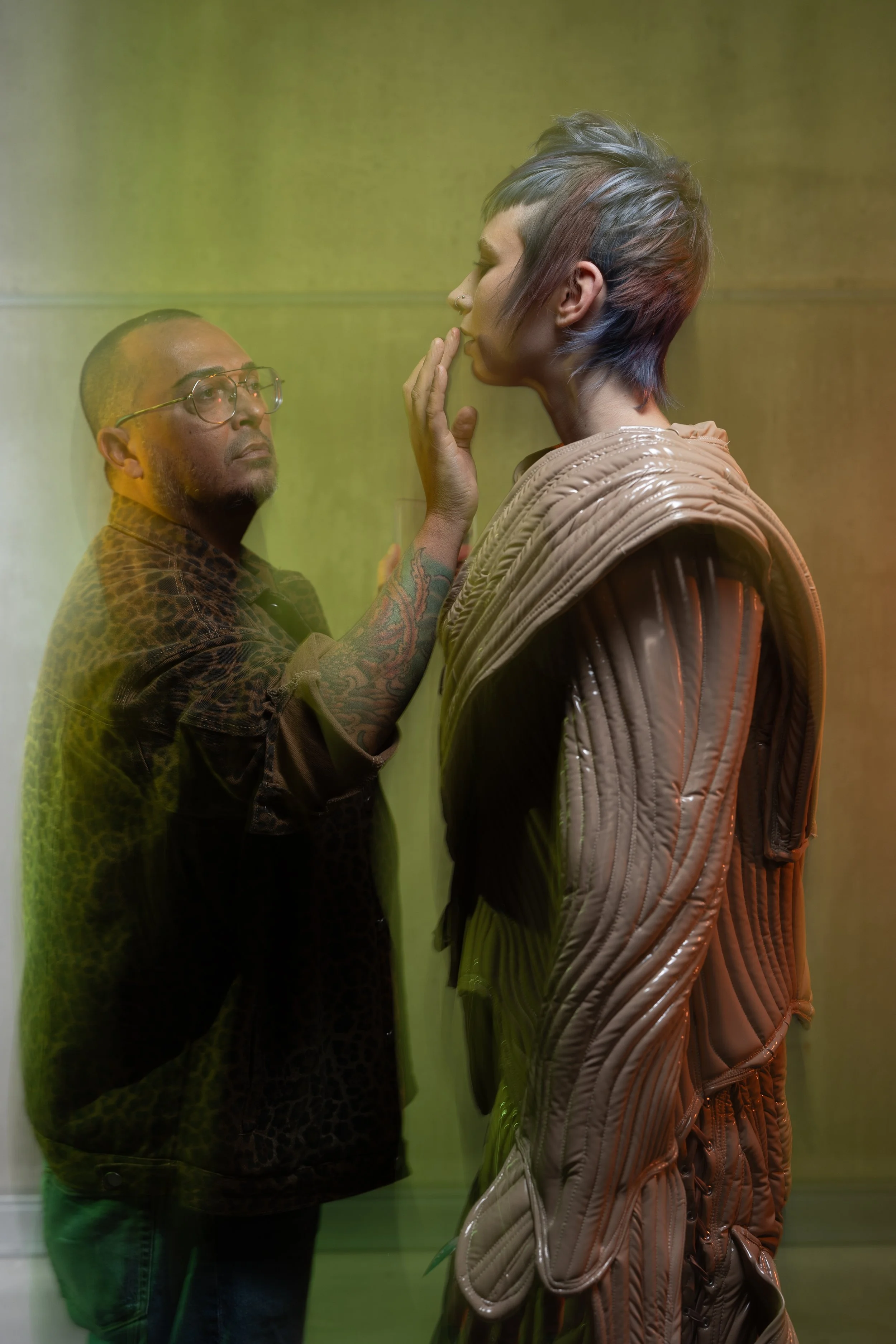Make-up artist touching up a model in the middle of the shoot. One person, with a shaved head, glasses, and tattoos, is touching the face of the other person, who has short, multi-colored hair and is dressed in a textured outfit.