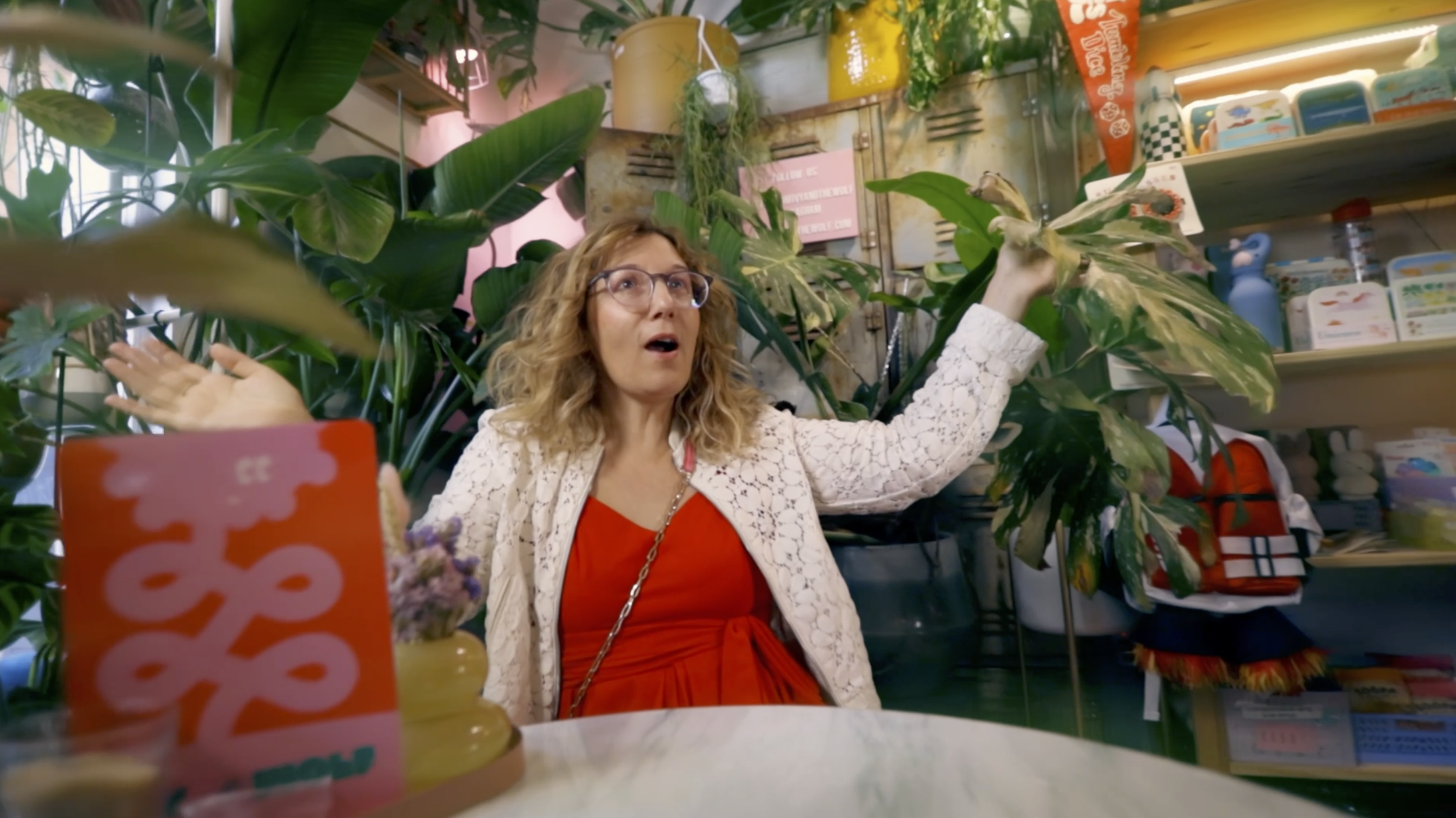 A woman with curly blonde hair and glasses is sitting at a table in a plant and gift shop. She is holding a plant with large leaves in one hand and gesturing with the other. The shop has green plants and various colorful gift items and products on shelves behind her.