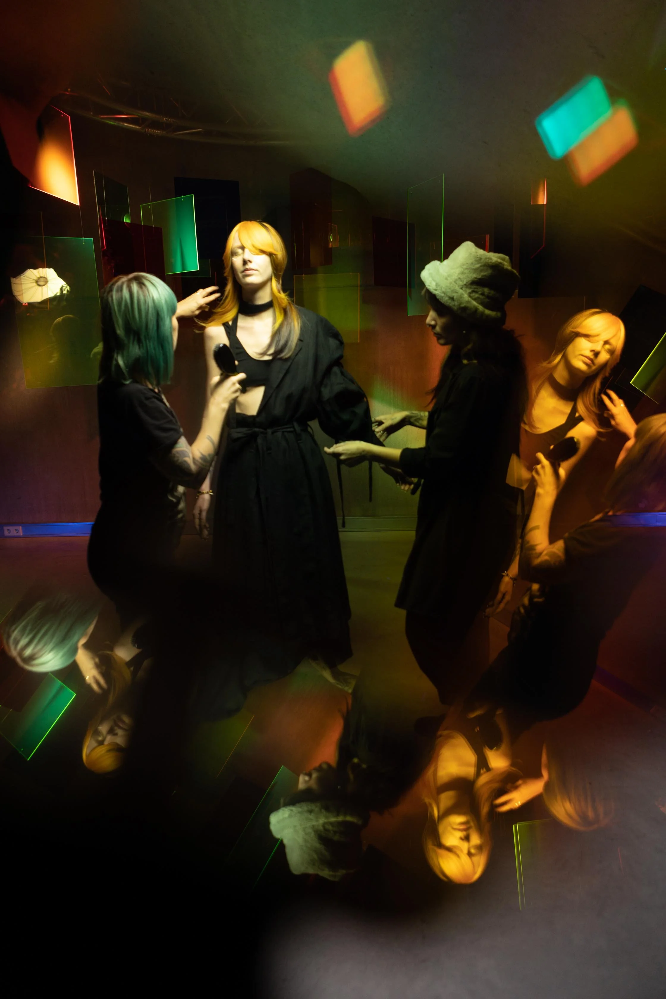 Four women with dyed hair and tattoos, one being interviewed with a mirror and flashlight, are in a room with colorful reflective panels and a dark background. The scene shows a mix of flat and reflected images.