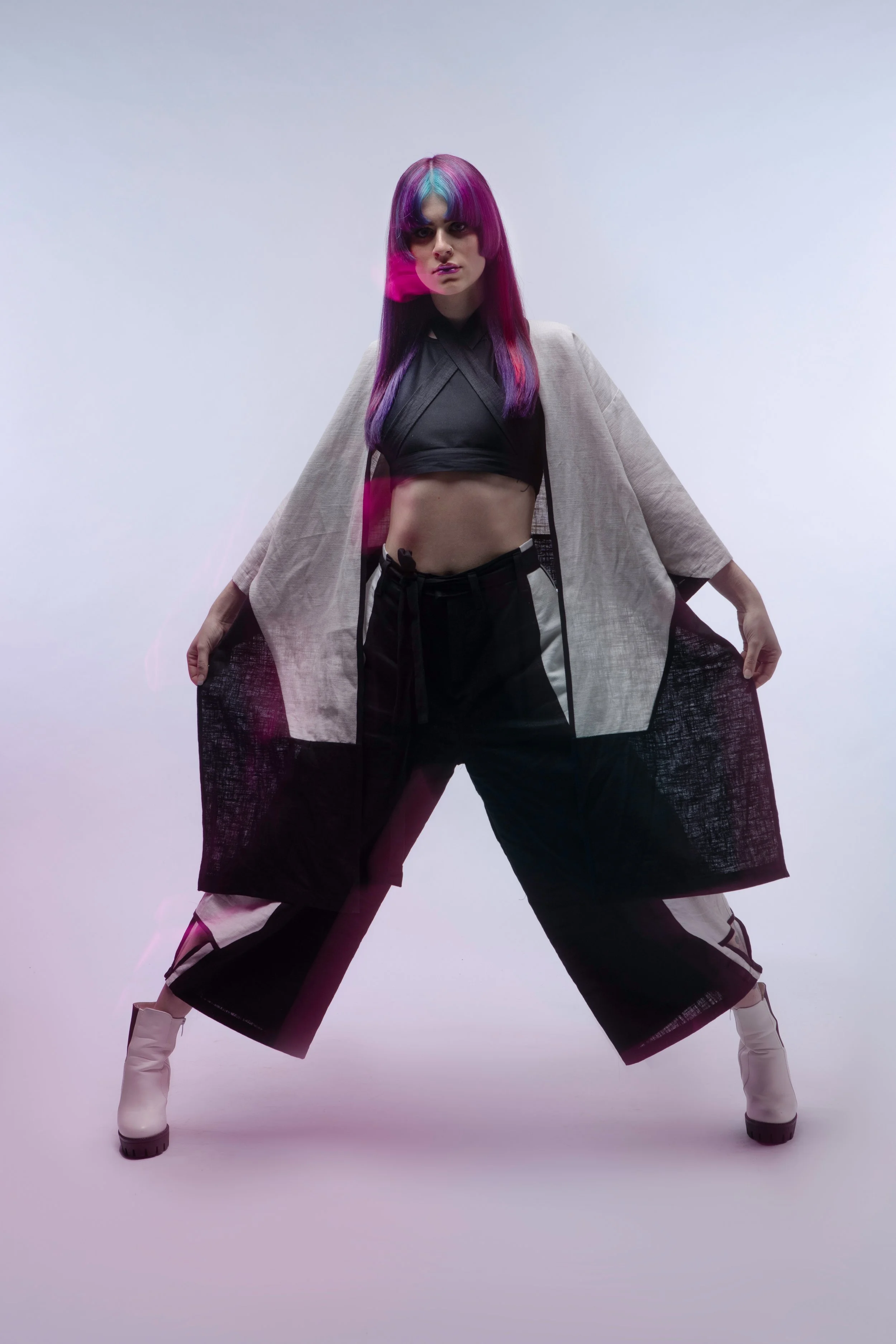 A woman with long, colorful hair styled with shades of purple, pink, teal, and blue, posing with legs apart and hands holding coat open. She wears a black crop top, baggy black cargo pants with white details, beige ankle boots, and a beige coat with 