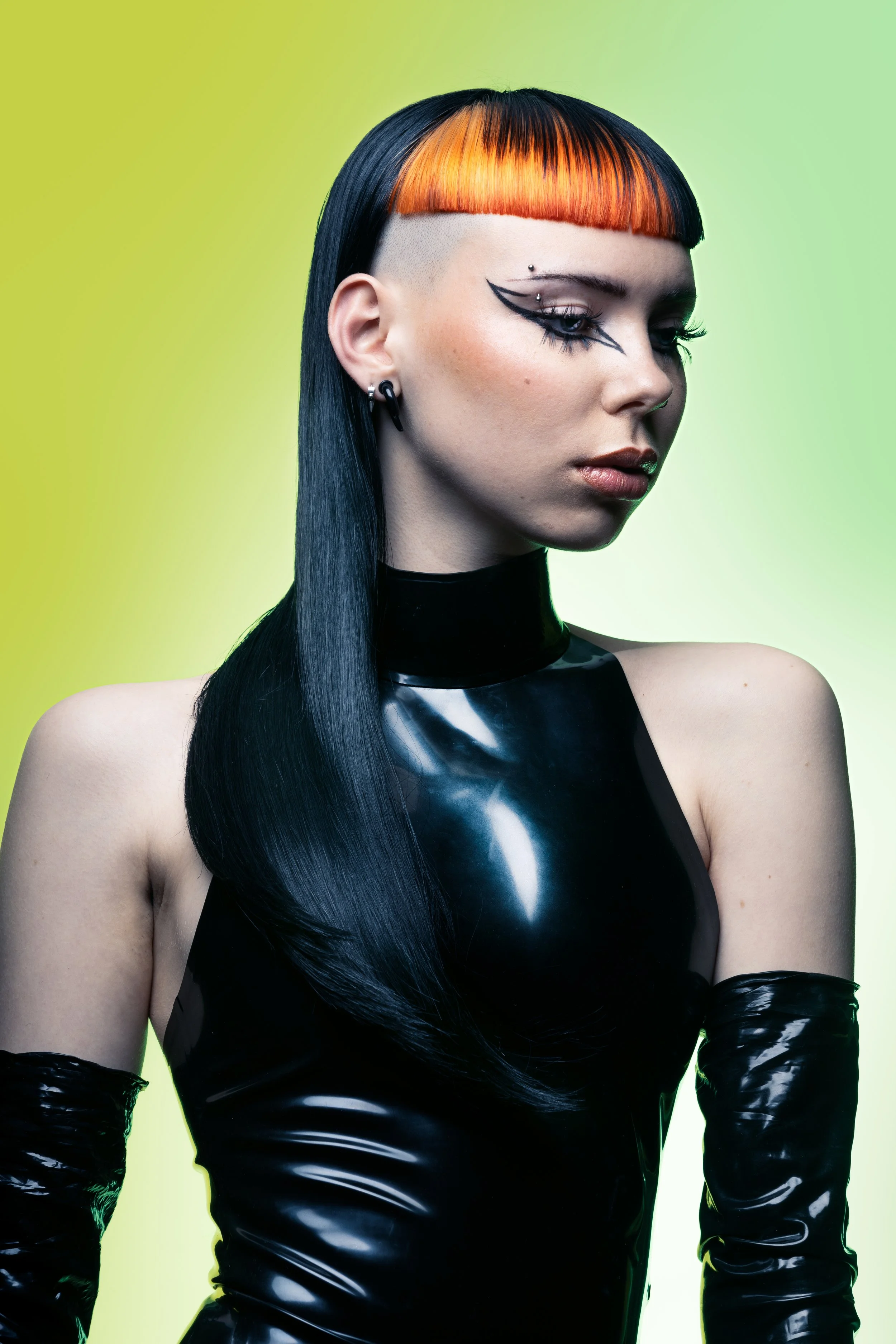 A woman with straight black hair and orange bangs, wearing black latex gloves and a matching black latex top, poses against a gradient green background.