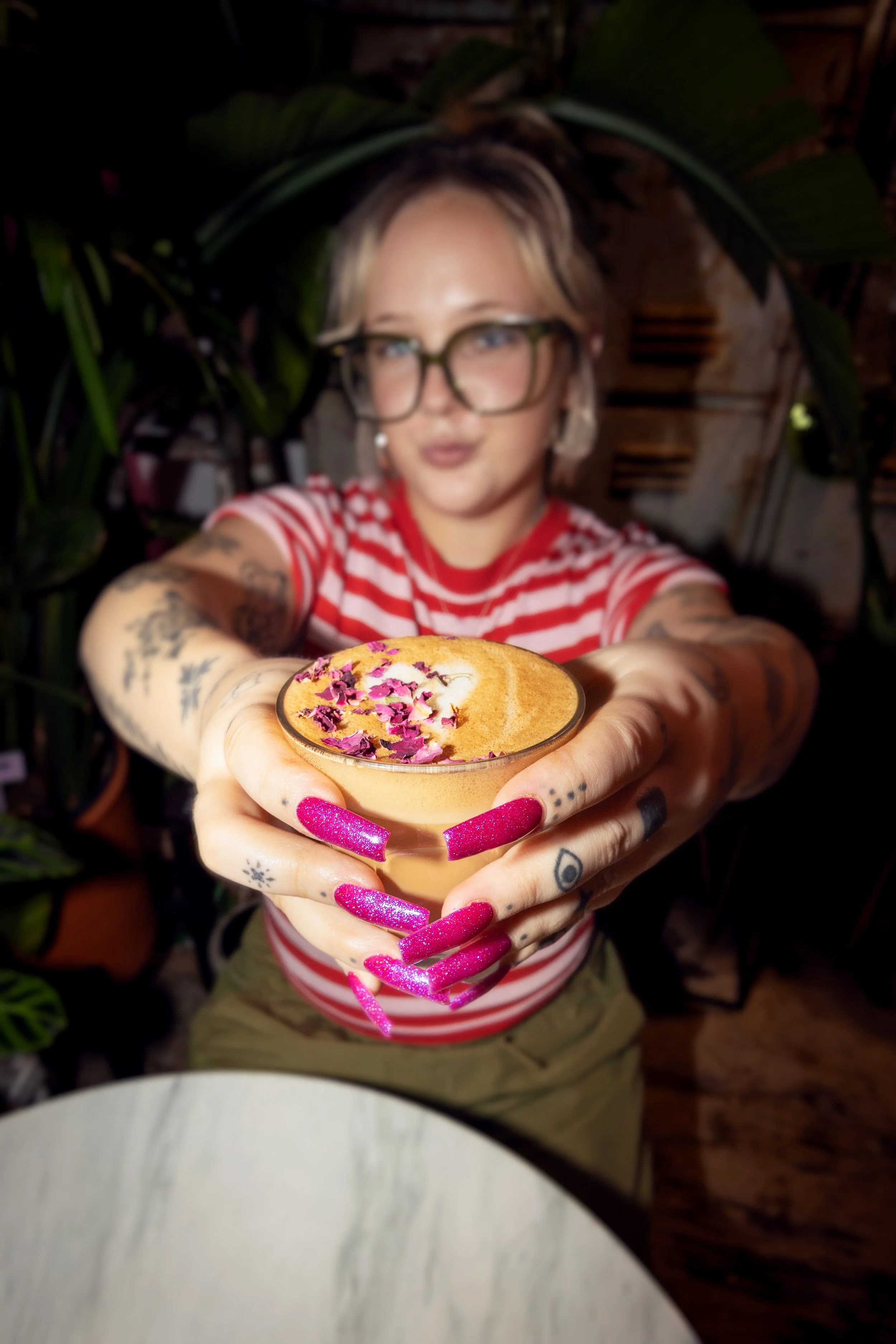 A woman with tattoos and pink glitter nails holding a latte with rose petals on top, extending it toward the camera.