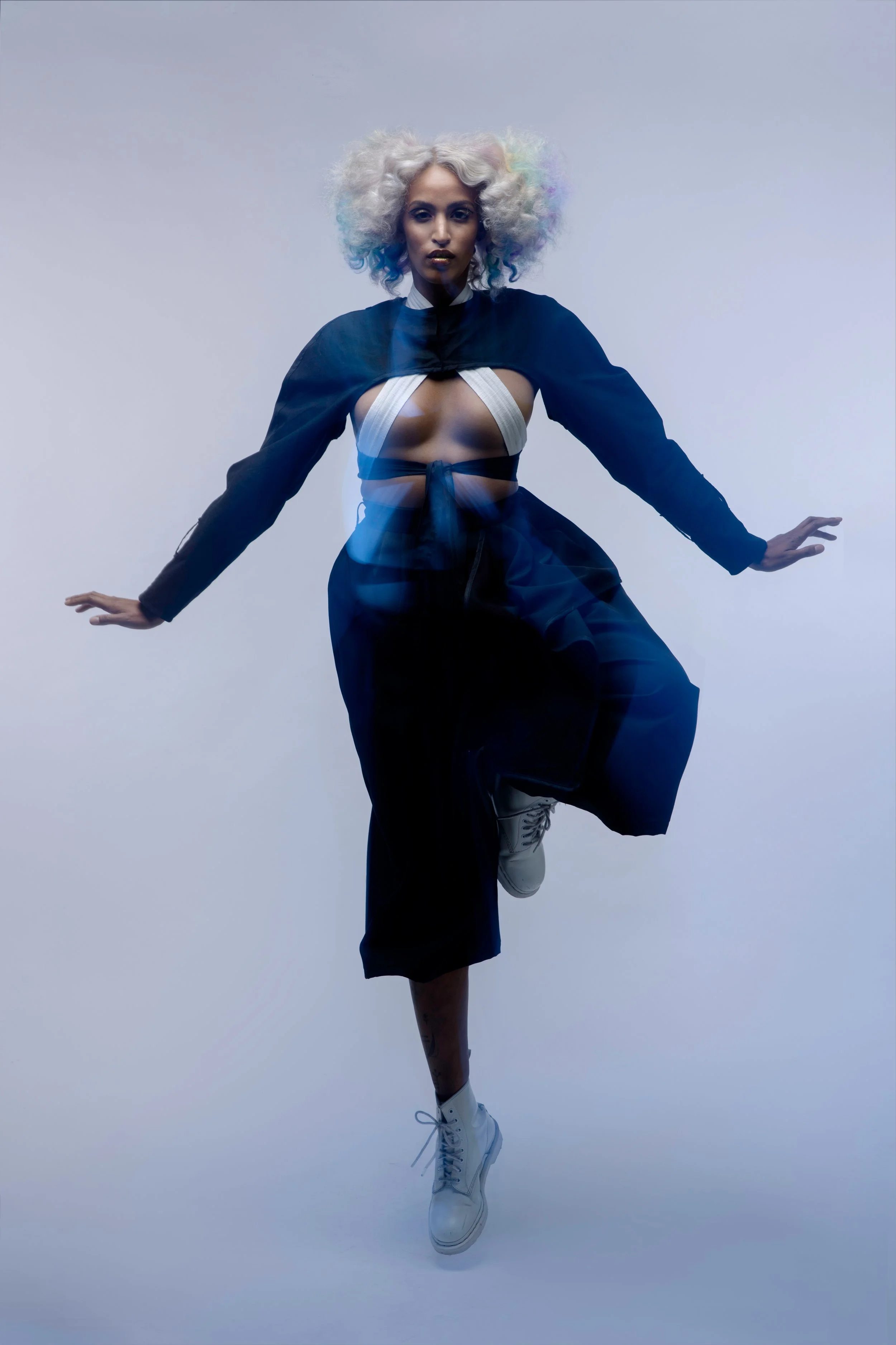 A person with colorful, curly hair and dark makeup poses against a plain background, wearing layered and stylish clothing including a black jacket, beige bandeau top, black pants, and white boots.