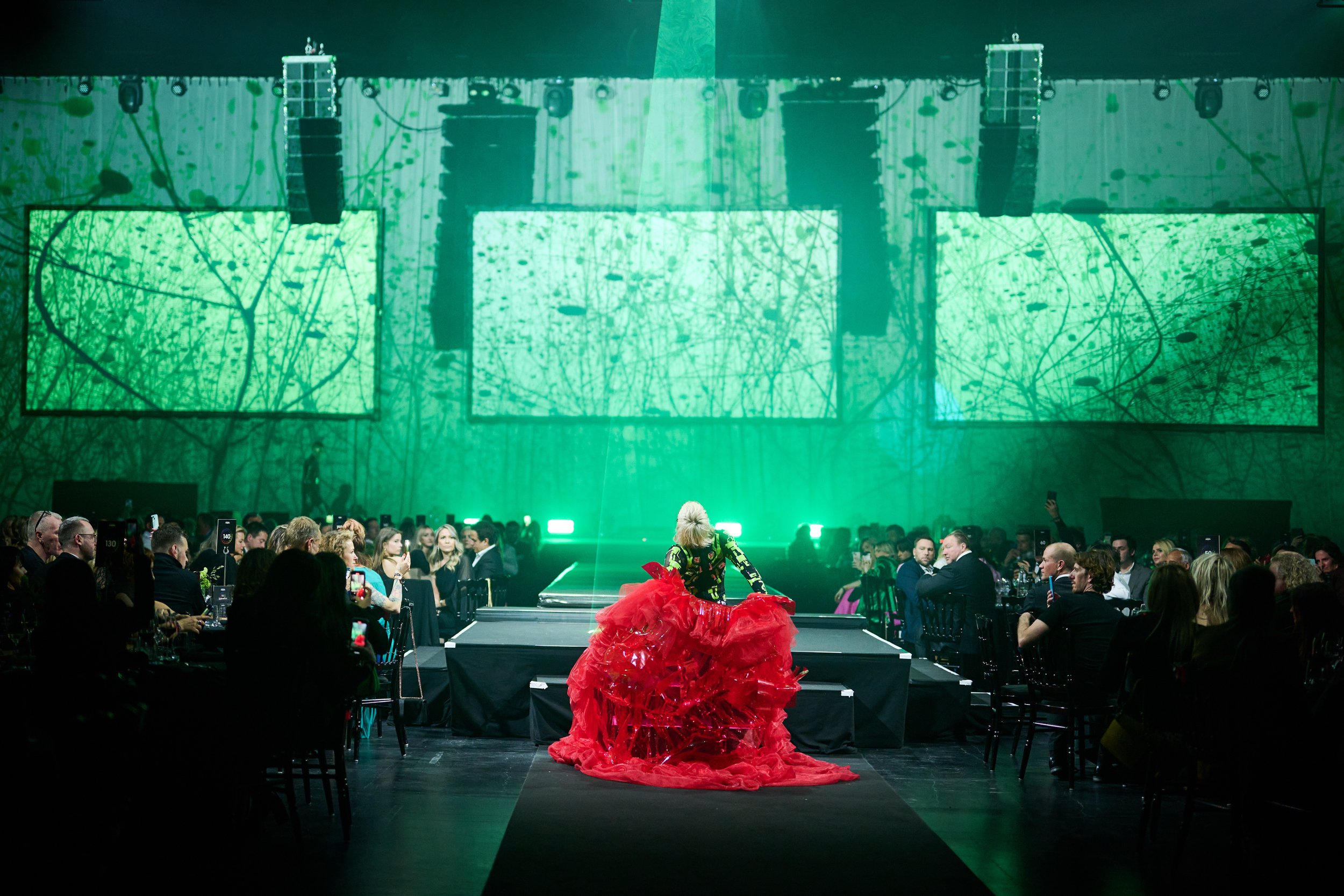 A performer on stage wearing a colorful top and a dramatic red tulle skirt, facing away from the audience in a dark venue with green lighting and large screens displaying abstract patterns, surrounded by seated guests.