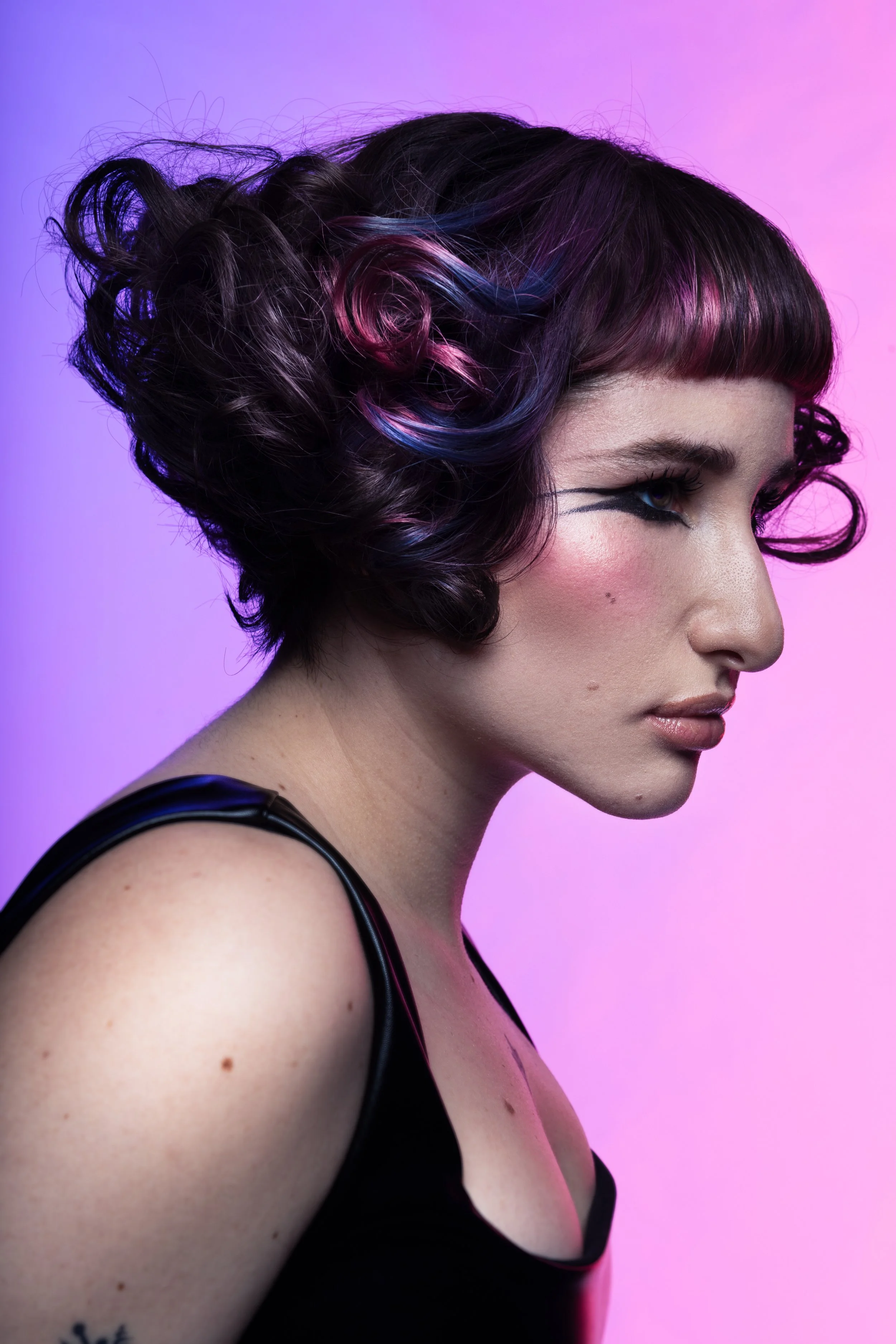 A young woman with short, curly hair dyed with shades of pink and blue, wearing a black top, against a pink and purple gradient background.