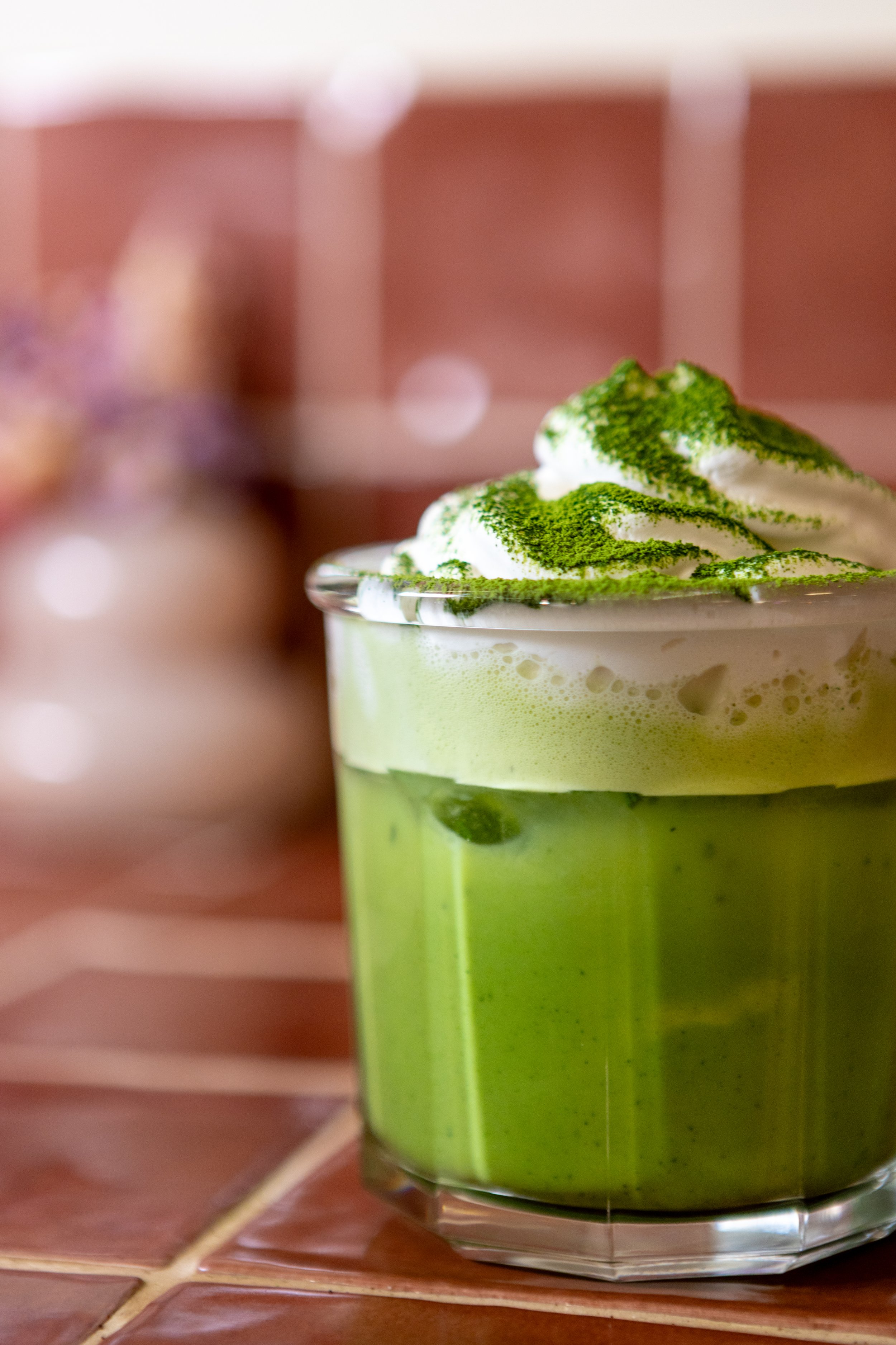 A glass of green matcha latte topped with whipped cream and sprinkled with matcha powder, sitting on a tiled surface.