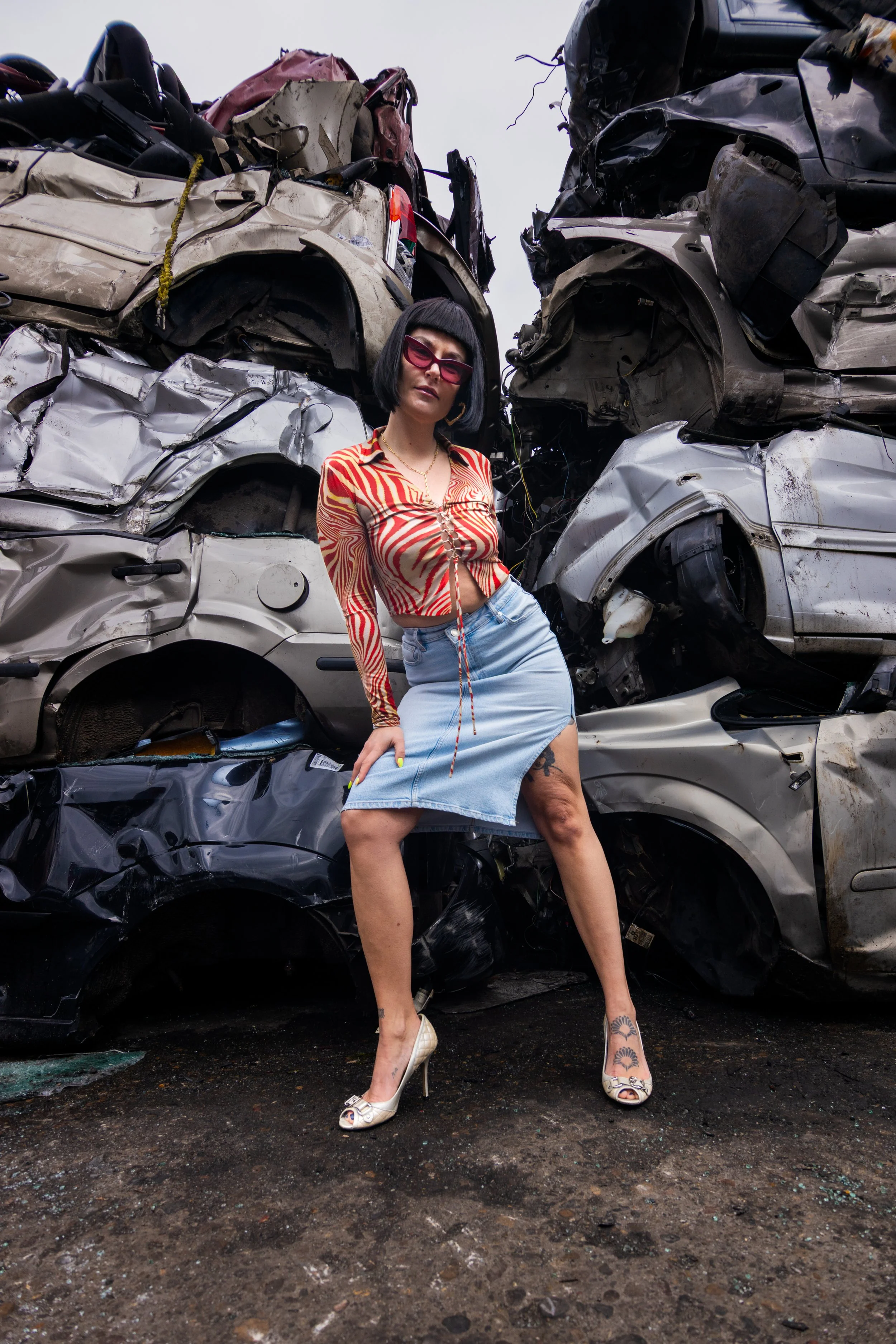 A woman standing in front of a pile of crushed, wrecked cars, wearing sunglasses, a colorful blouse, a denim skirt, and high heels.