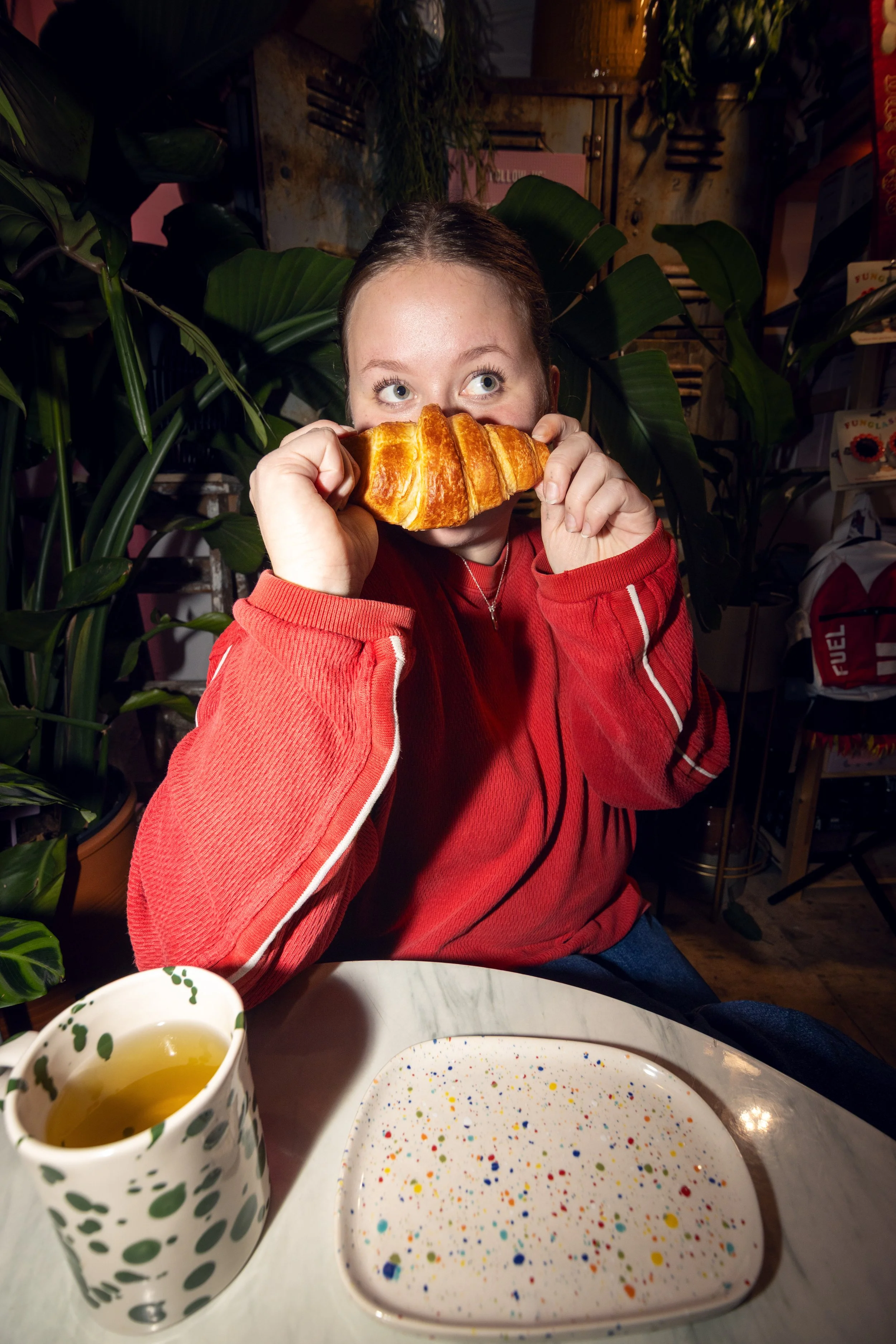 A young woman in a red jacket sitting at a table, holding a croissant near her face, with a colorful speckled plate and a mug with green spots in front of her, surrounded by green plants in a cozy indoor setting.