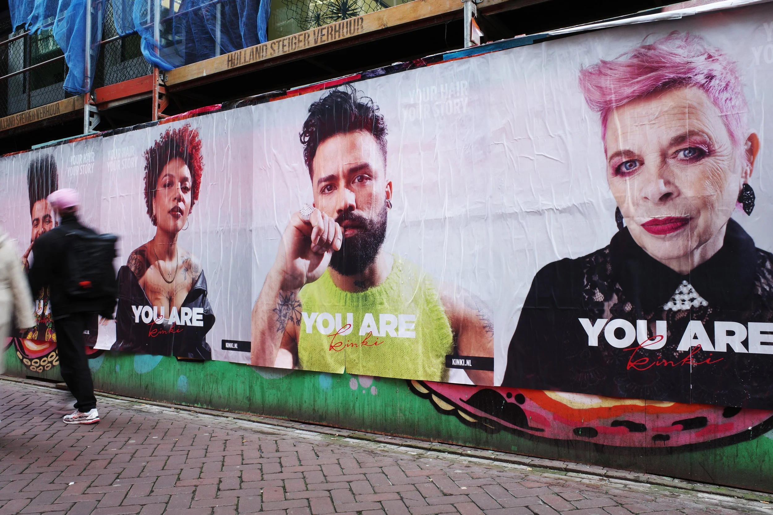 Large city street billboard displaying portraits of diverse individuals with the text 'YOU ARE' and the name 'Kink' in red. People walking by on a brick sidewalk.
