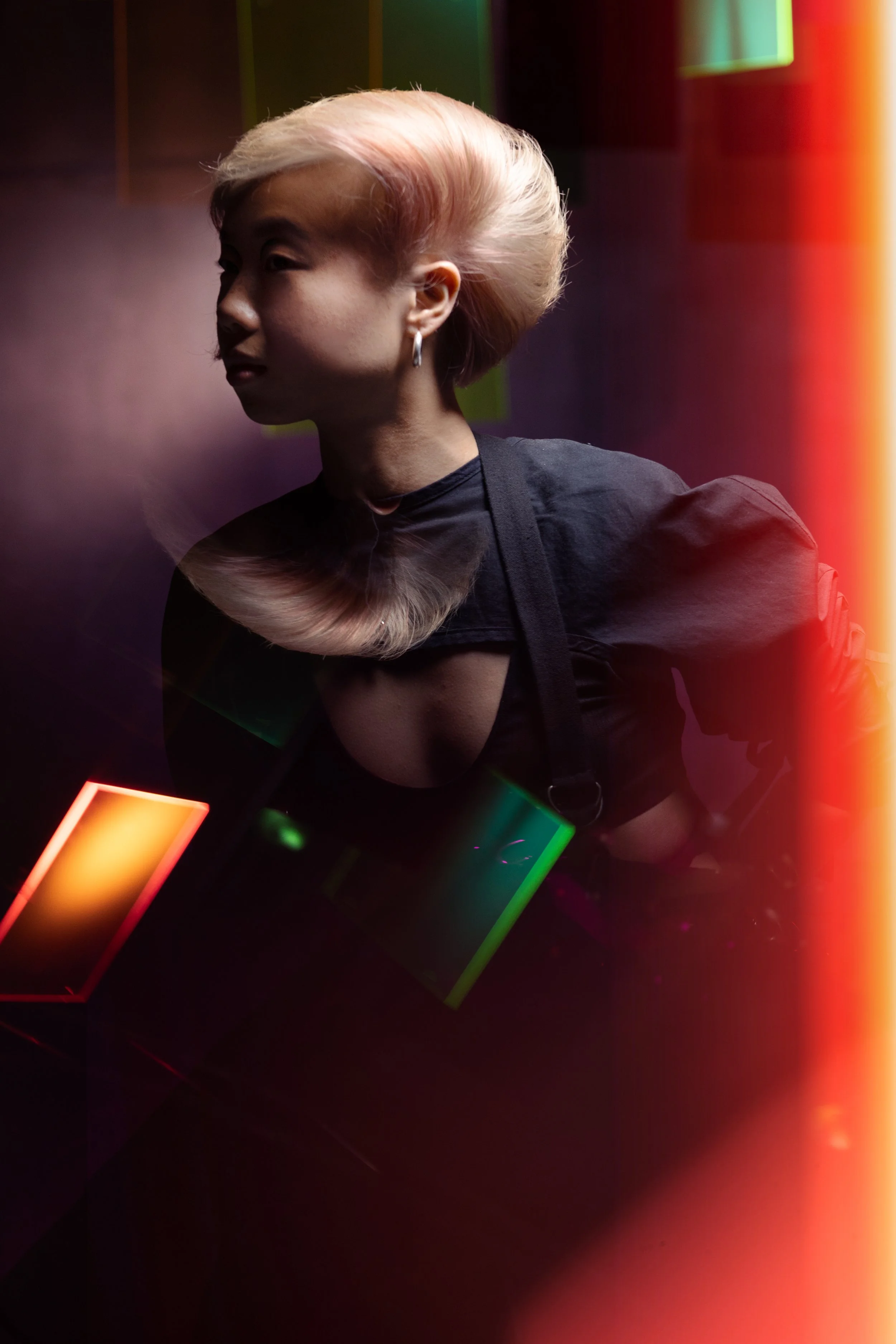 A woman with platinum blonde hair styled in an elaborate updo, wearing a black top with a plunging neckline and earrings, in a dark setting with multi-colored neon lights and light streaks.