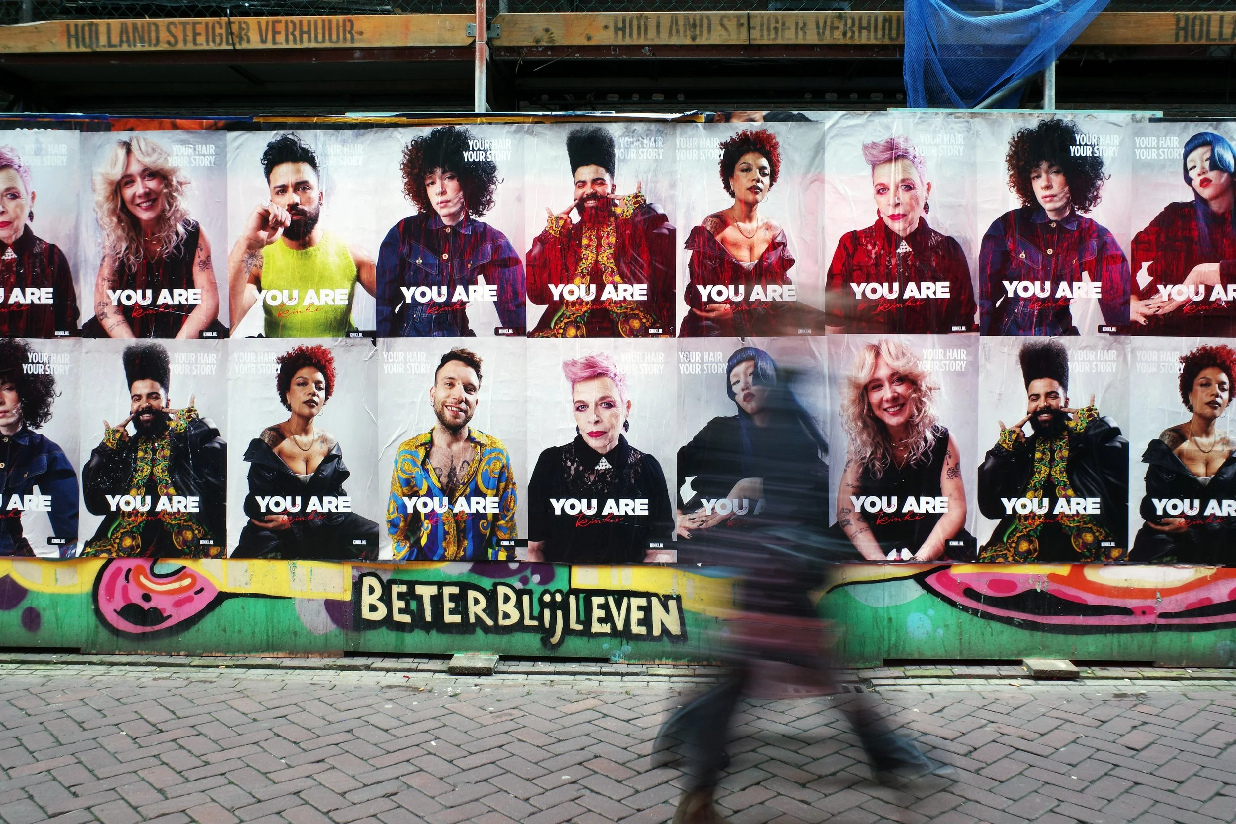 Street view with a colorful graffiti wall and posters of diverse people, with the phrase 'BETTER BE LOVE' on the wall, and a person walking past in motion blur.