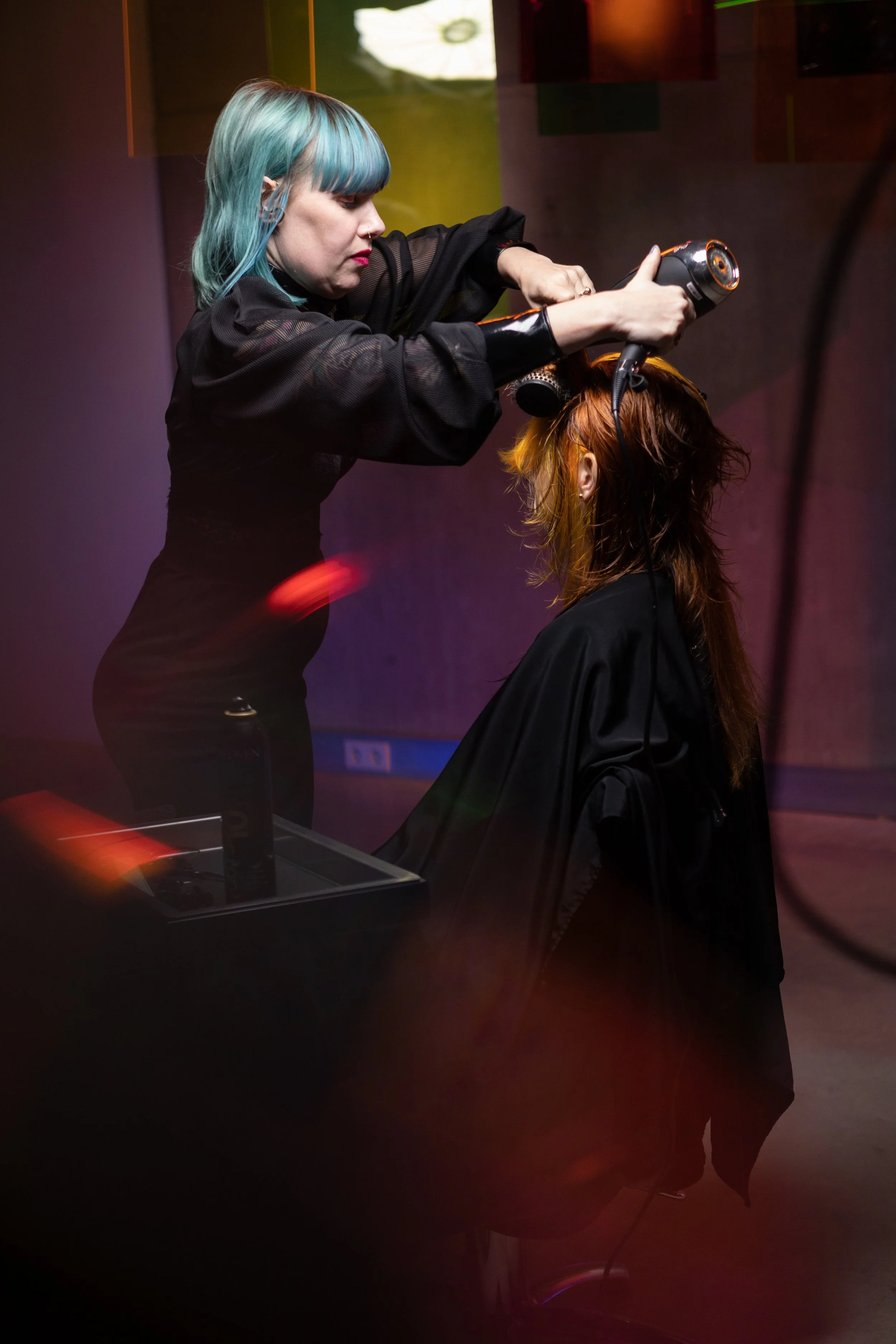 A hairstylist with teal hair blow-drying a client's hair in a salon with colorful lighting.