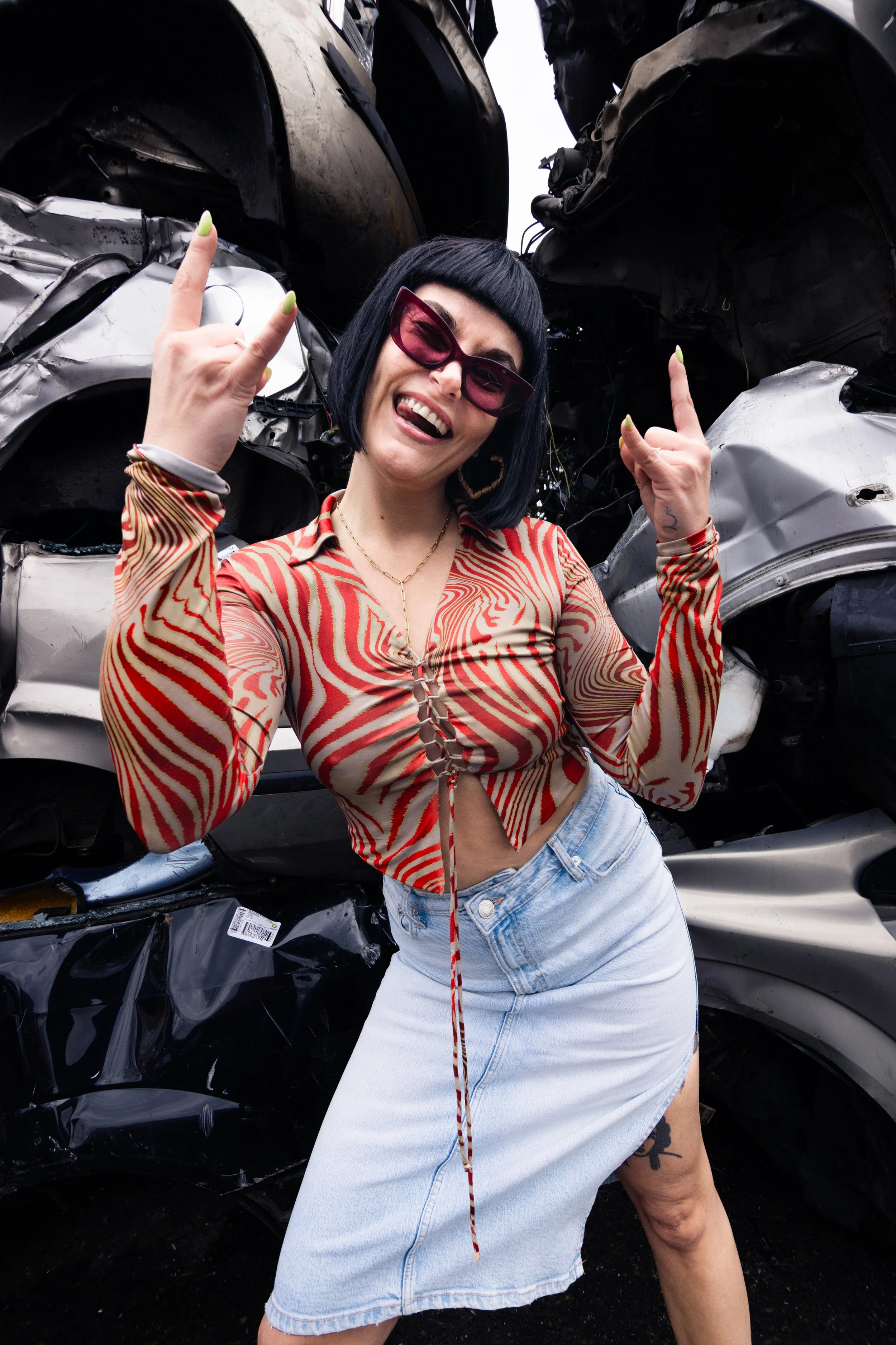Woman with black bob haircut, sunglasses, and bold makeup smiling and giving rock and roll hand gestures in front of a pile of crushed cars, wearing a patterned long-sleeve top and a light denim skirt.