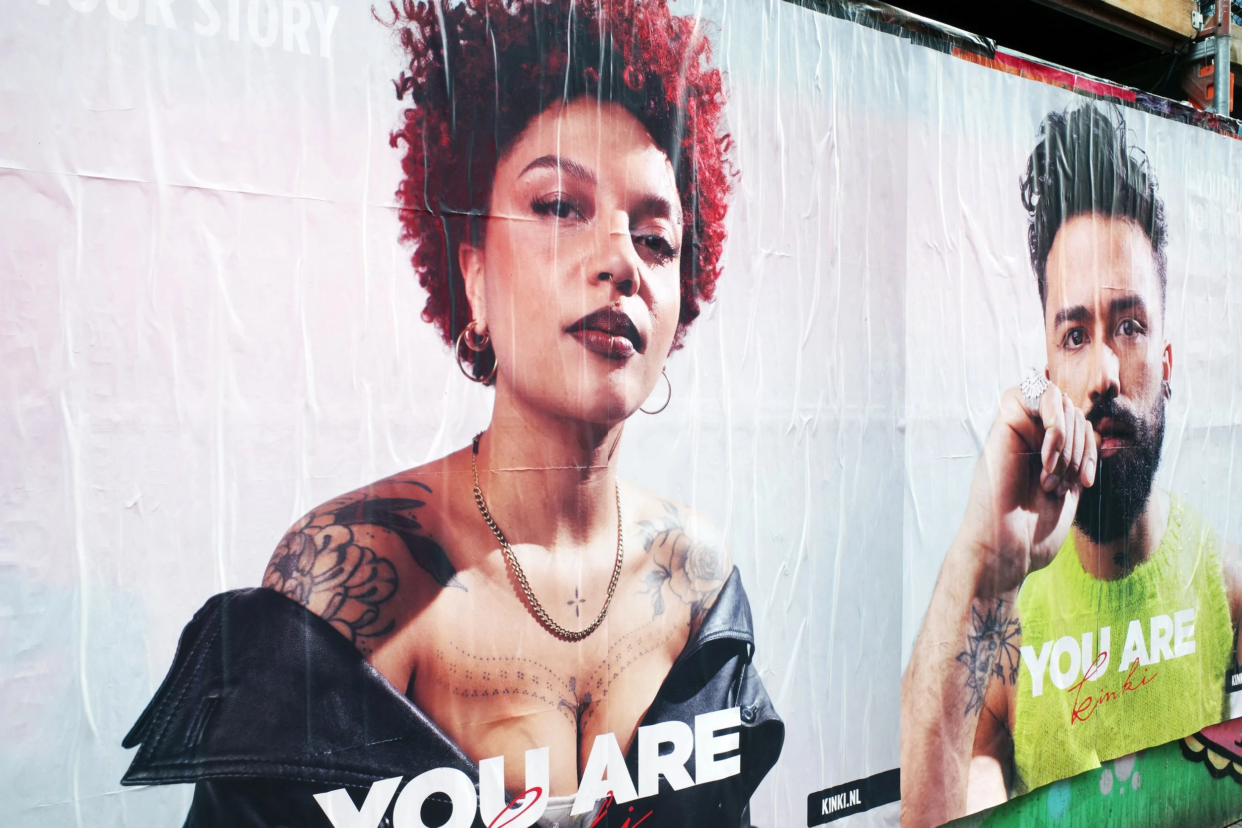 Large outdoor billboard featuring two people, a woman with red curly hair and tattoos on her shoulders and chest, and a man with a beard and black hair. The woman is wearing a black off-shoulder jacket and jewelry, and the man is holding a ring. Both
