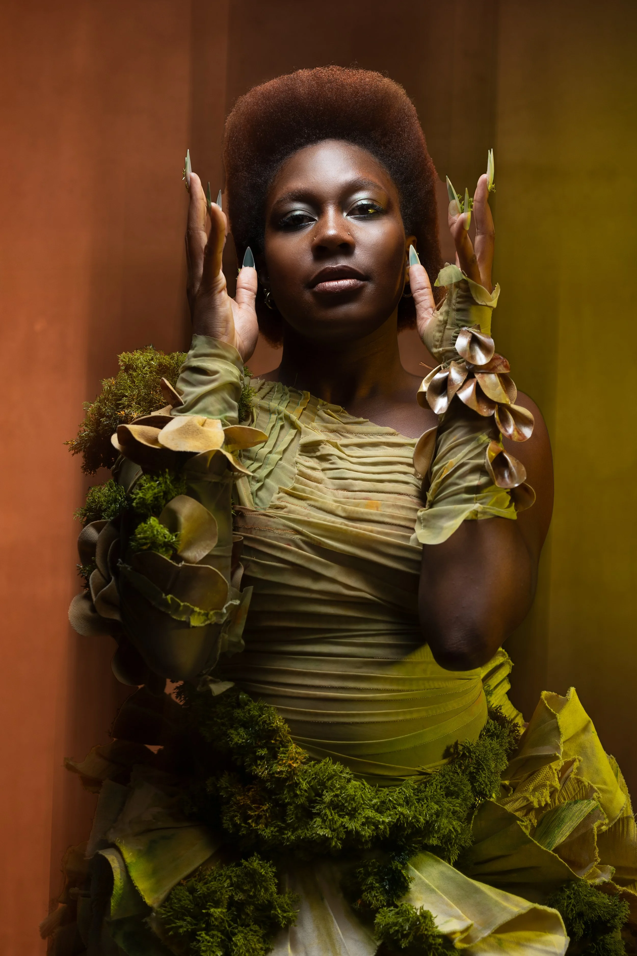 A woman with natural hair styled in an afro, wearing a green, nature-inspired outfit adorned with leaves and moss, posing with hands near her face.