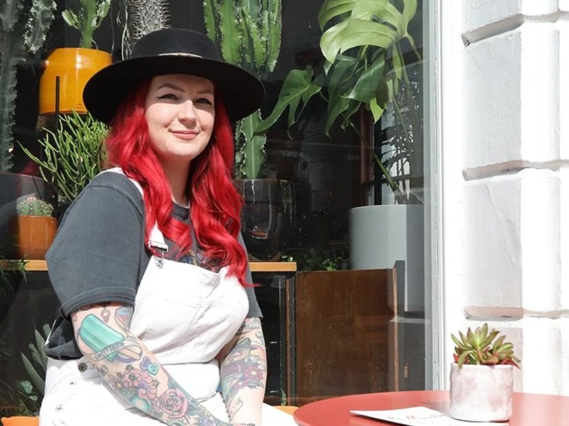 Woman with red hair wearing a black hat, gray t-shirt, and white apron, sitting inside near a display of potted plants.