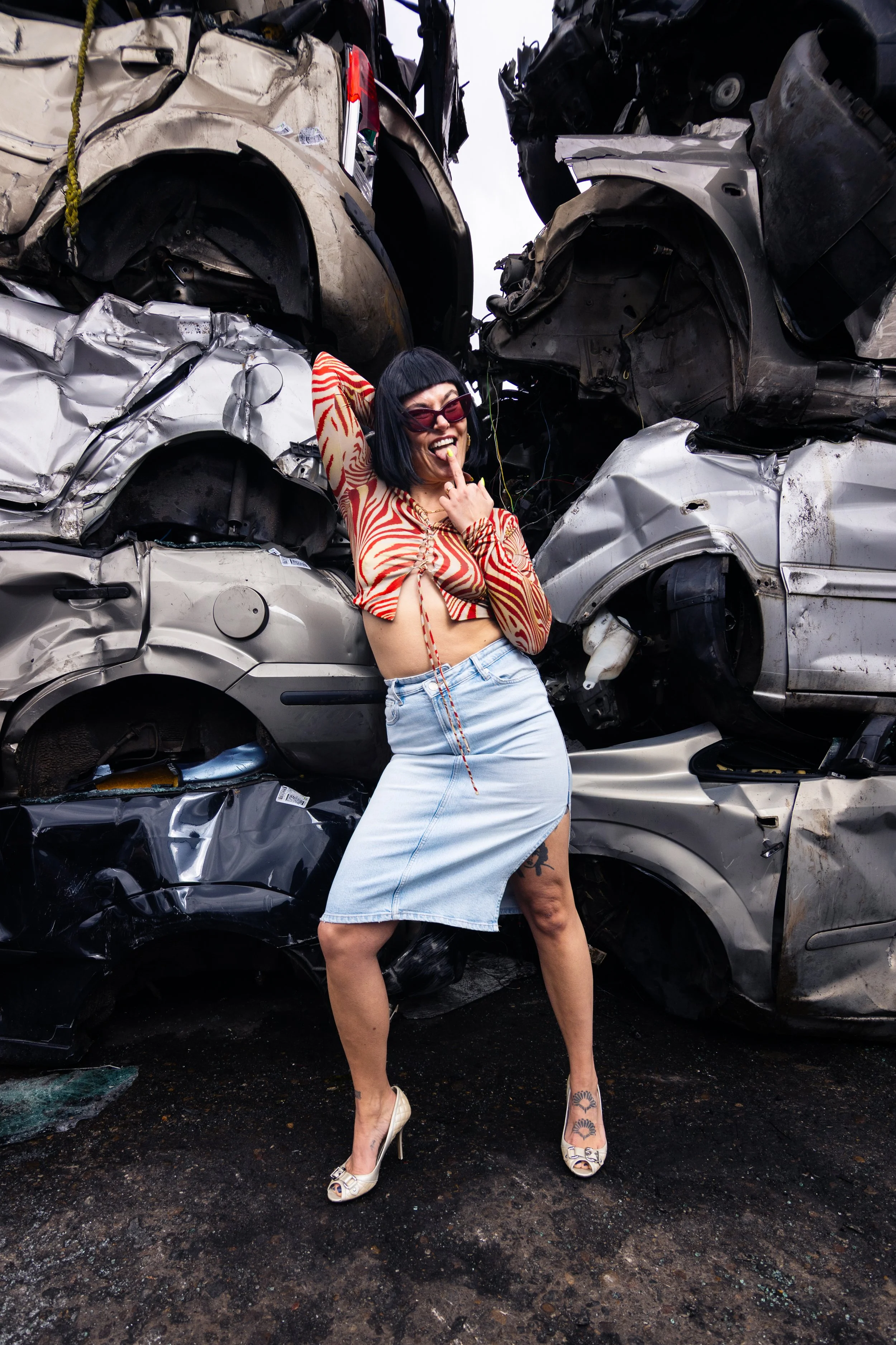 Woman with black bob haircut, sunglasses, and tattoos posing in front of stacked crushed cars, making a playful gesture with her mouth open and one finger pointing to her face.