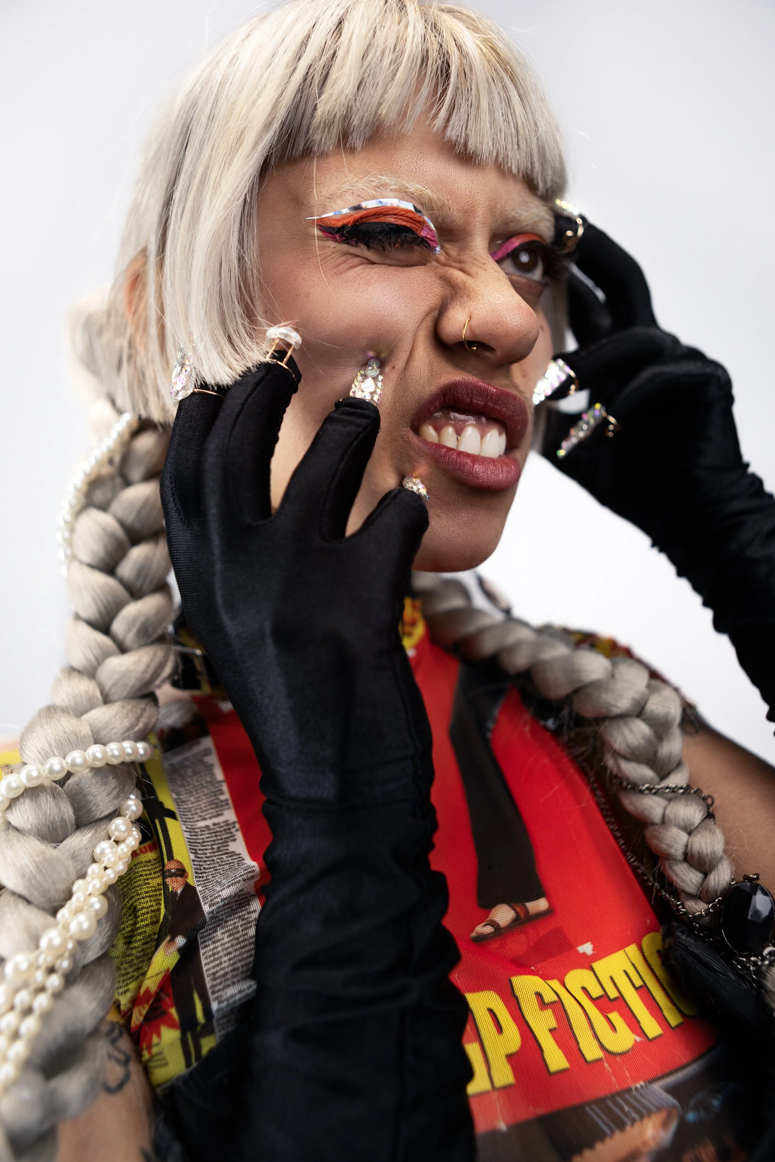 A person with platinum blonde hair in braids, wearing black gloves, bold makeup with colorful eye shadow and winged eyeliner, and a t-shirt with a graphic print. They are touching their face with a fierce expression.