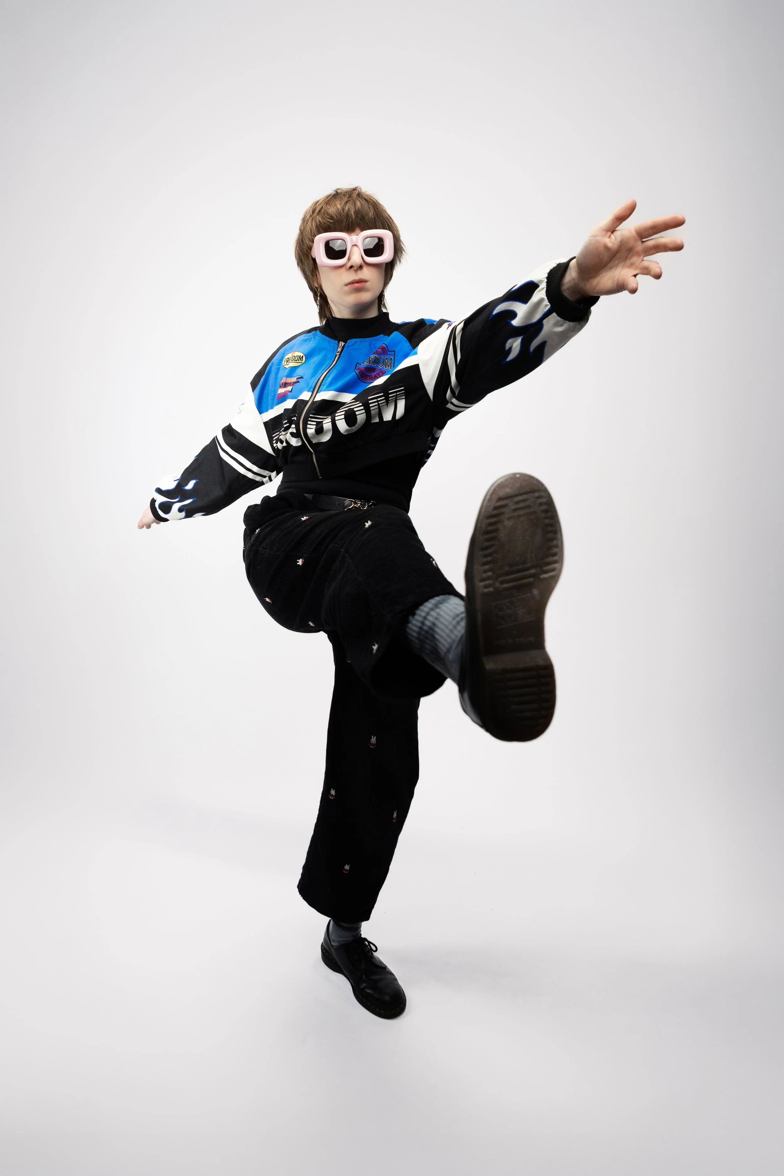 Person wearing a blue and black jacket with patches, black pants with small designs, and pink sunglasses, kicking one leg forward in a dynamic pose against a plain white background.