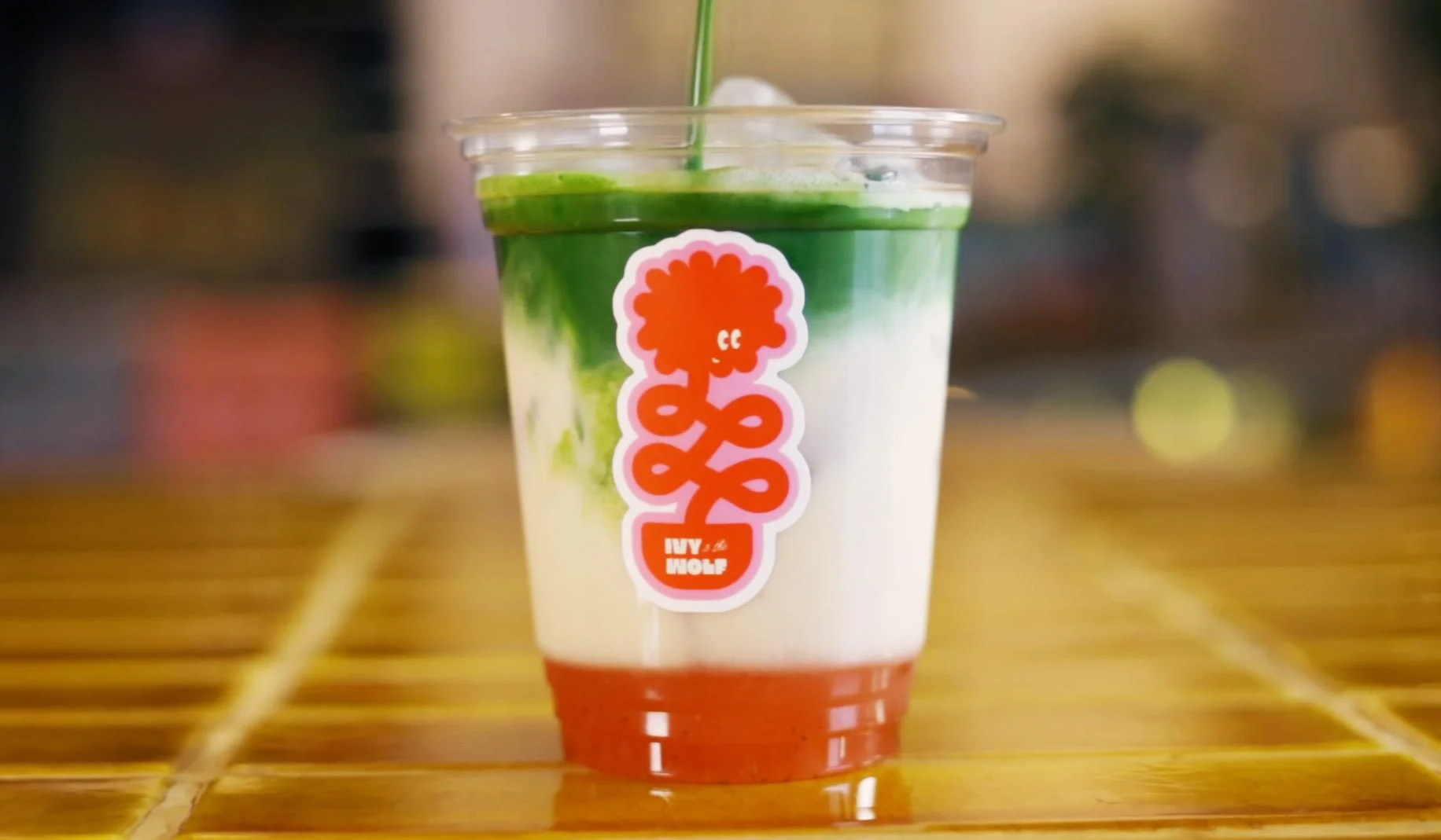 A clear plastic cup containing a layered drink with red at the bottom, white in the middle, and green at the top, with a green straw. The cup has a sticker with a red flower and spiral design, and the text "IVY WOLF."