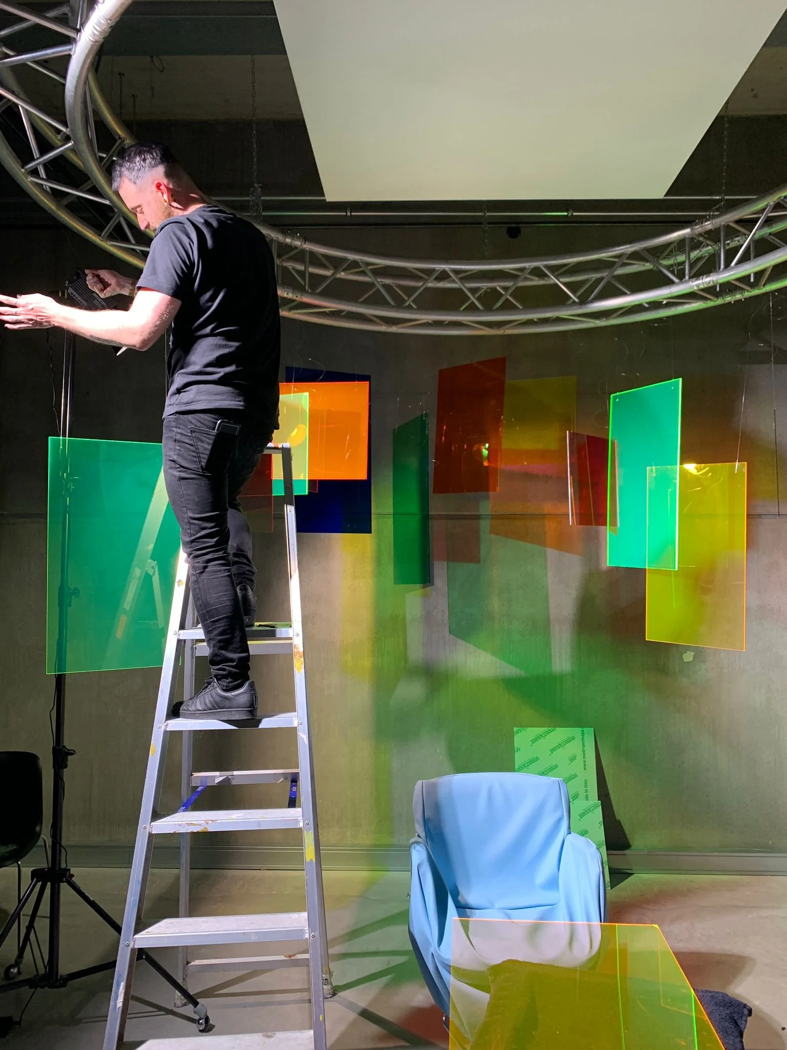 A person standing on a ladder working on hanging colorful acrylic sheets in a studio or exhibition space.