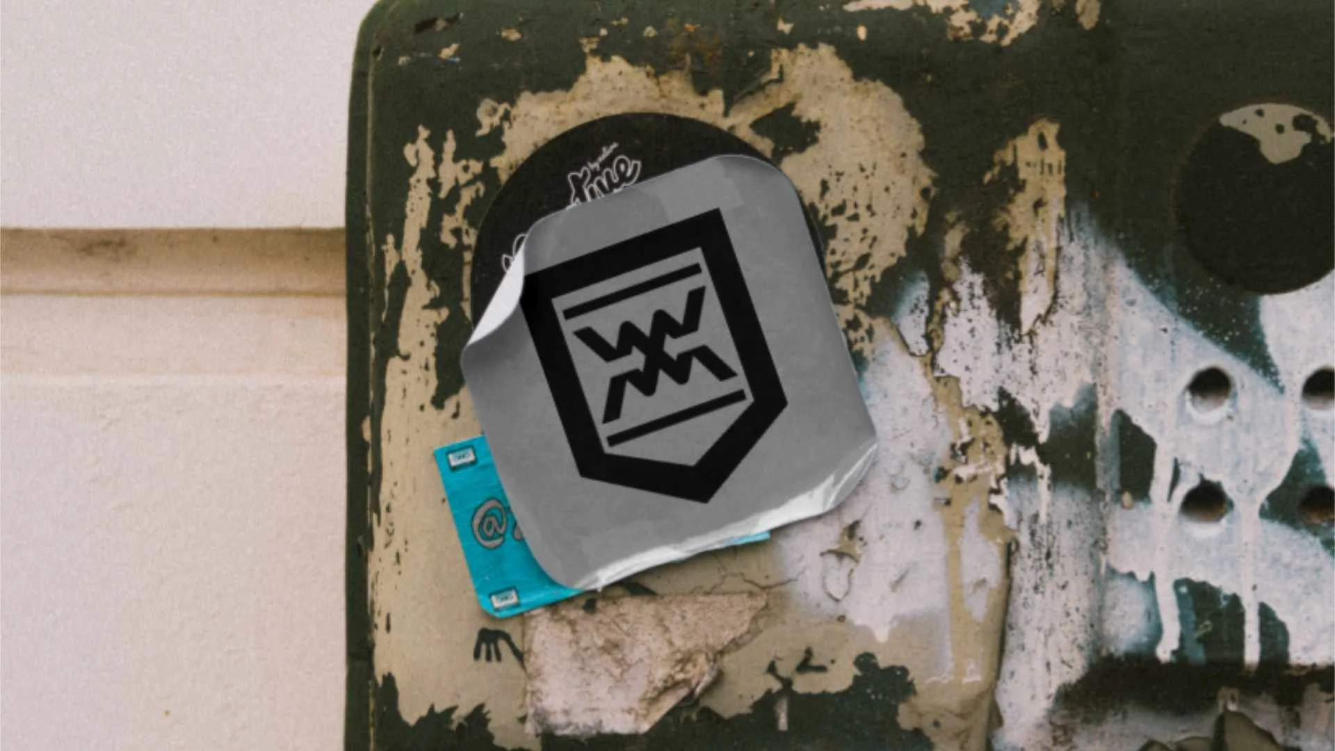 Close-up of a worn, black and beige metal surface with graffiti, peeling paint, and dirt, featuring a crumpled gray sticker with a black chemical equation symbol on it, partially covering a small blue sticker.