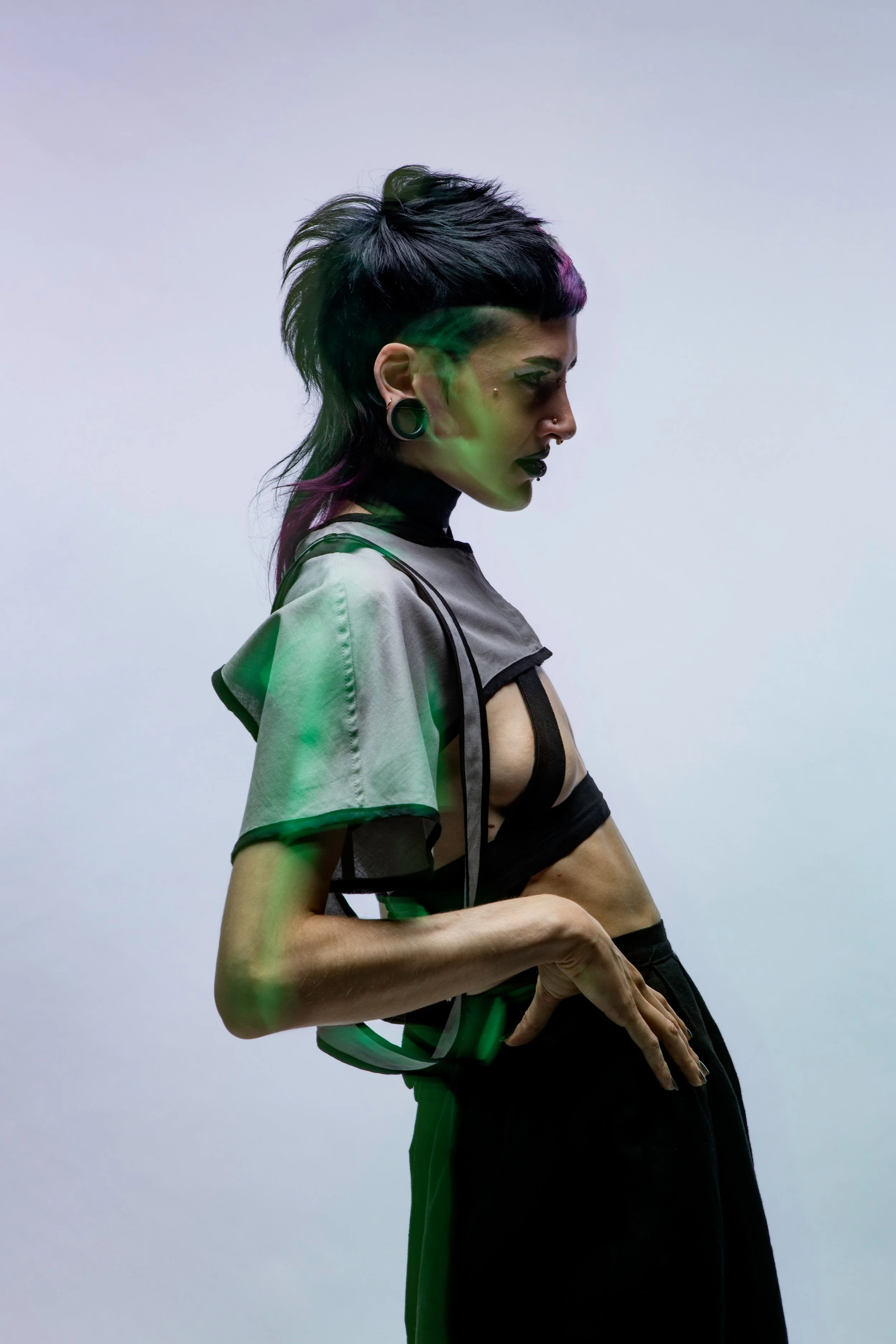 Side profile of a person with punk hairstyle, multiple facial piercings, and dark makeup, wearing a black and gray fashion outfit against a plain white background.