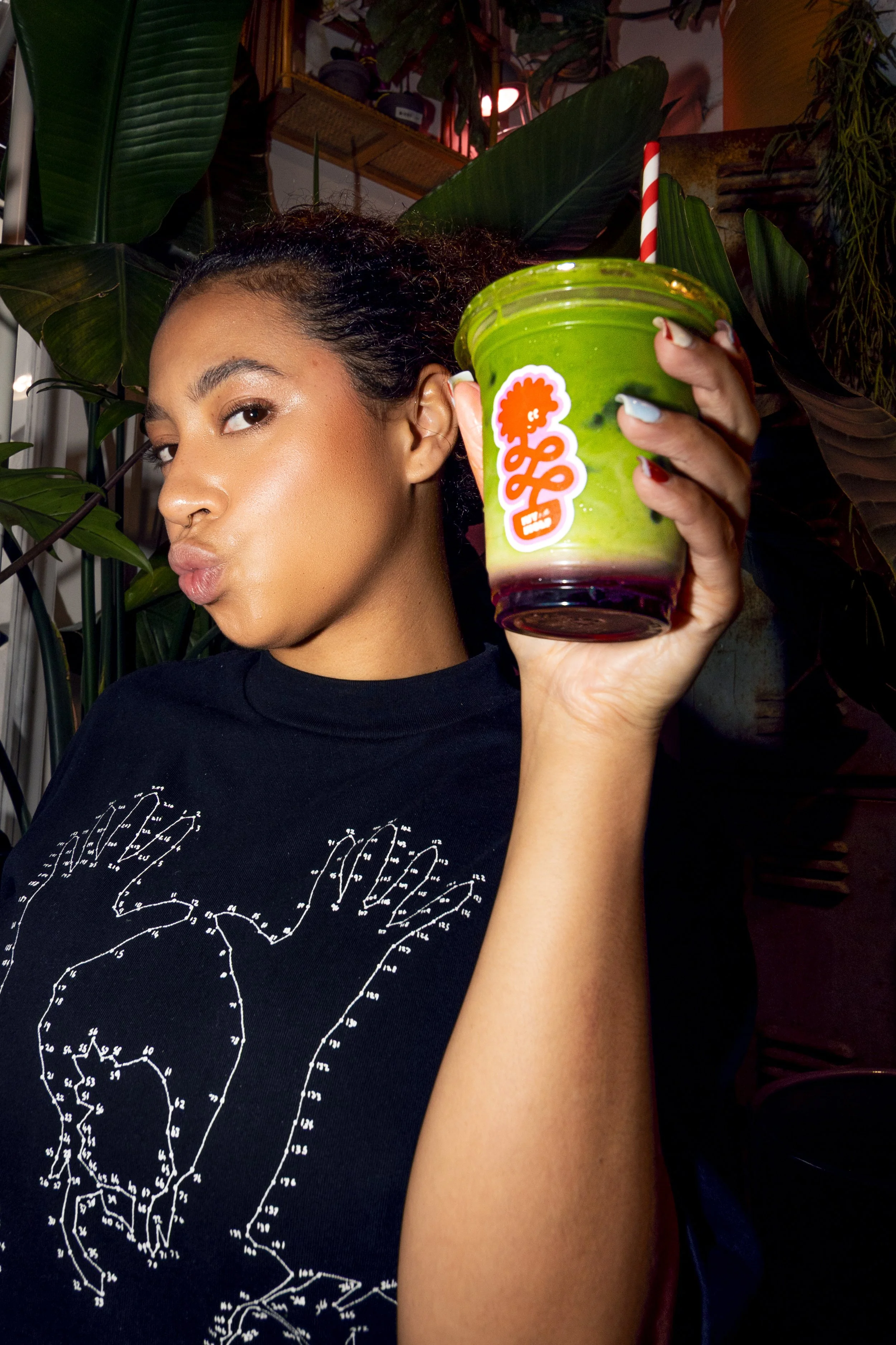 A woman holding a green and purple layered drink with a red and white striped straw in front of green plants, making a playful kiss face.