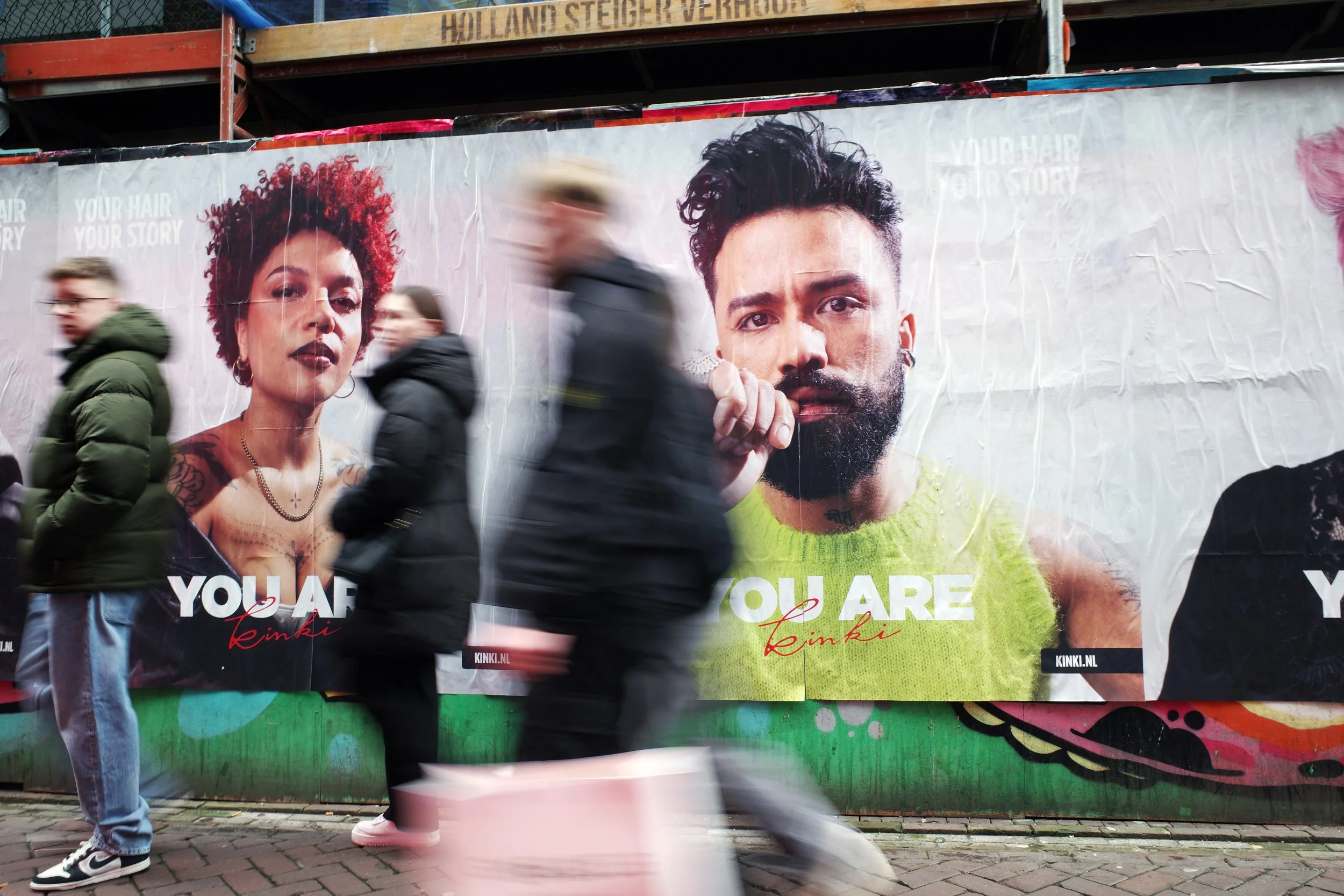 Pedestrians walking past large, colorful billboard advertisements featuring diverse models with the message "You Are Kinki."