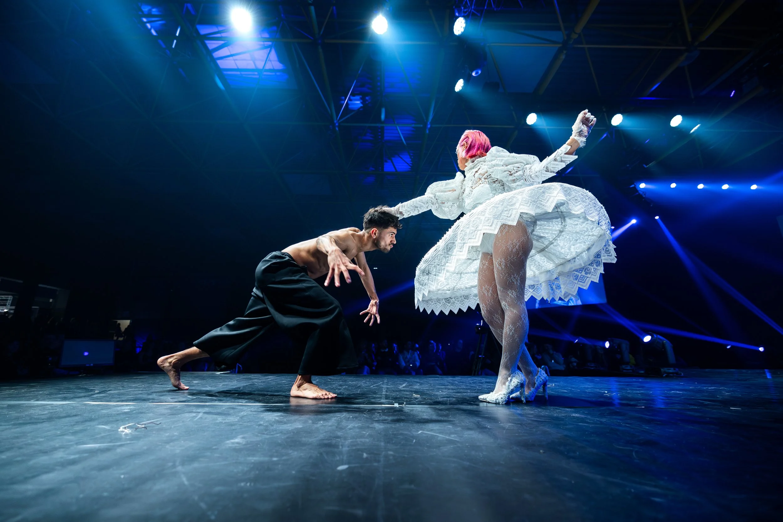 A male dancer with dark pants crouching and a female dancer with pink hair in a white lace dress raising her arm, performing on a stage with blue lights and a dark background.
