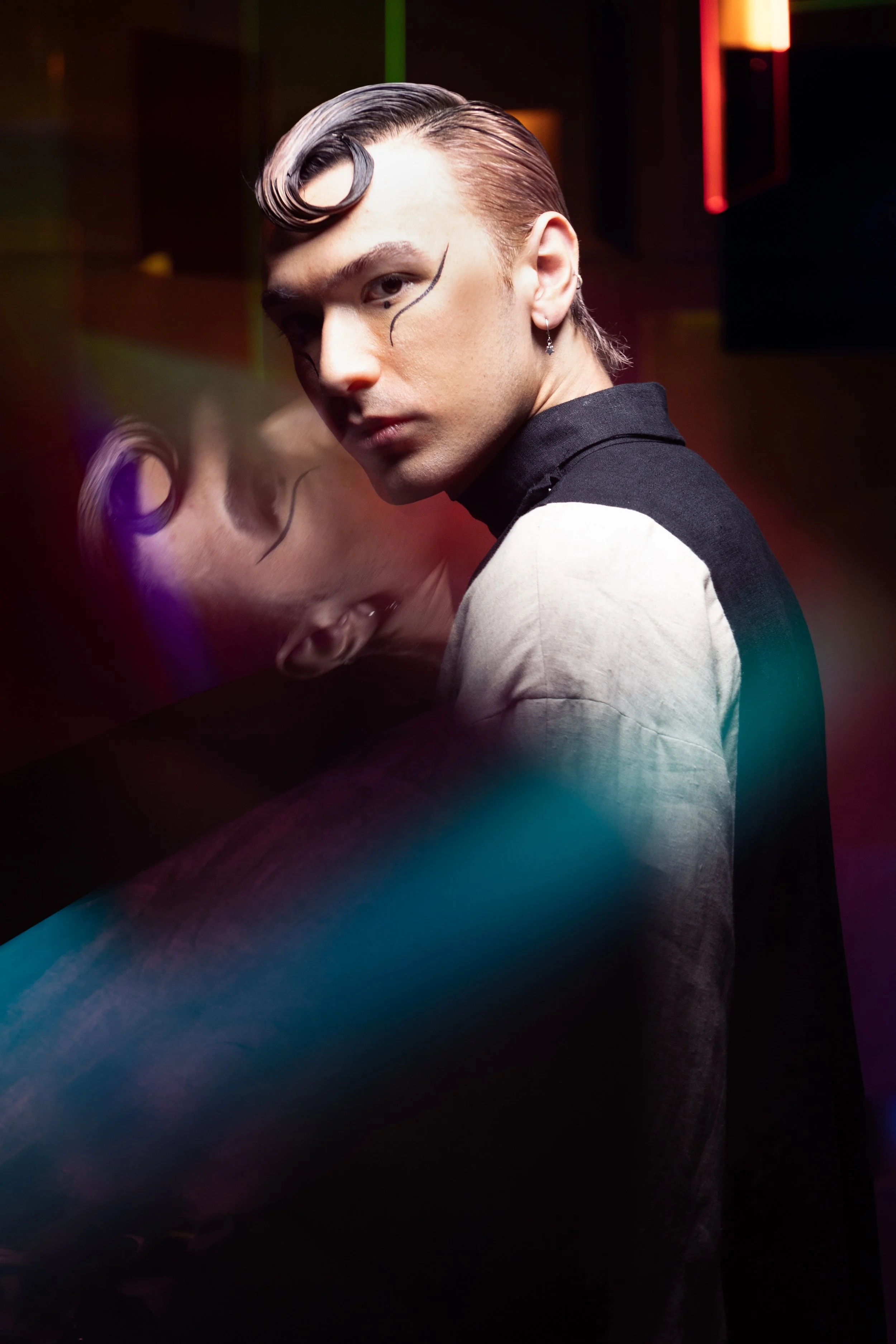 A young person with styled hair and makeup, wearing earrings, looking over their shoulder with a serious expression, with a colorful, blurred background.