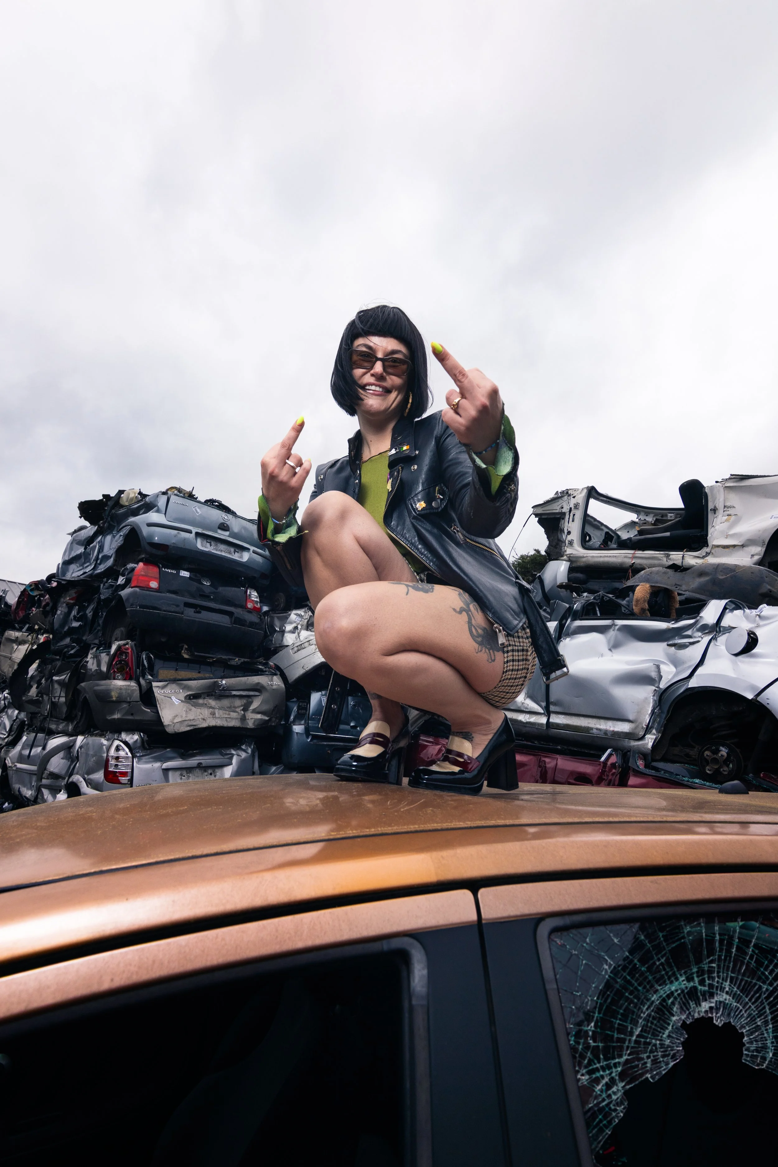 A woman with black hair, glasses, and tattoos squatting on top of a damaged car at a junkyard, making an offensive gesture with her middle fingers, smiling.