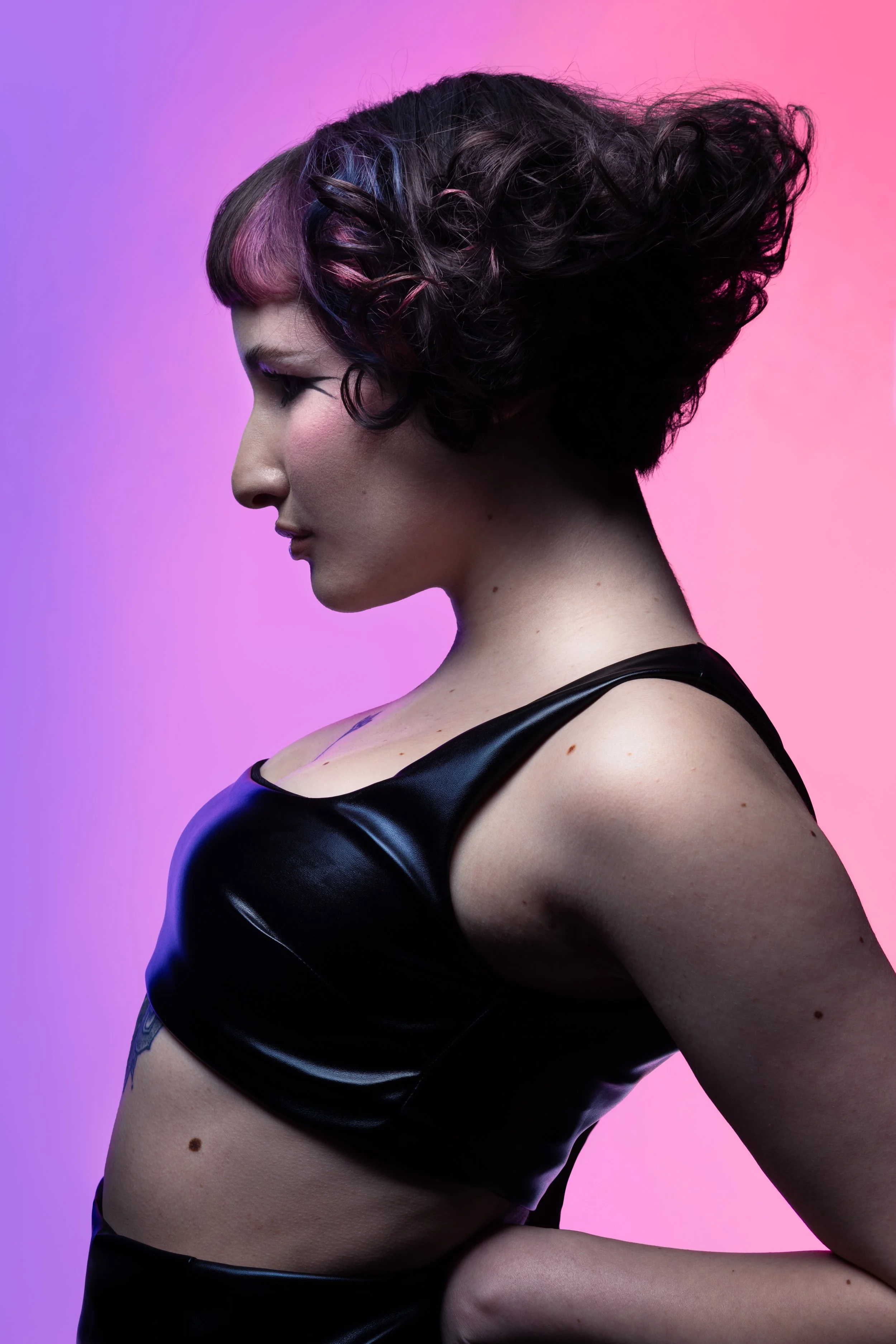 Side profile of a woman with colorful hair and makeup, wearing a black shiny top against a gradient pink and purple background.
