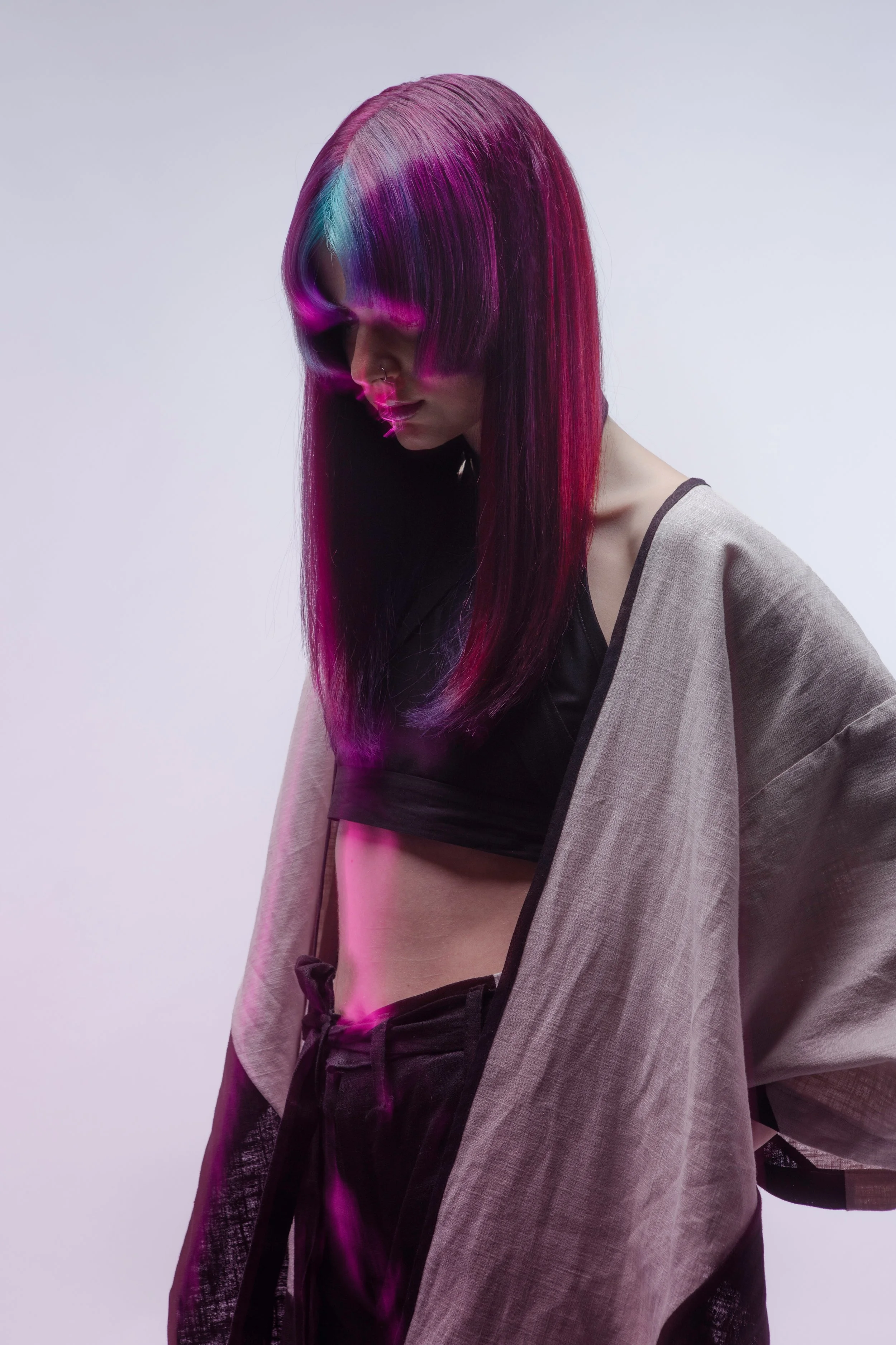 A young woman with long, straight hair that is dyed in shades of purple, pink, and blue, looking down with her eyes closed. She is wearing a black crop top, a beige oversized coat, and black pants, with pink lighting accentuating her hairstyle and ou