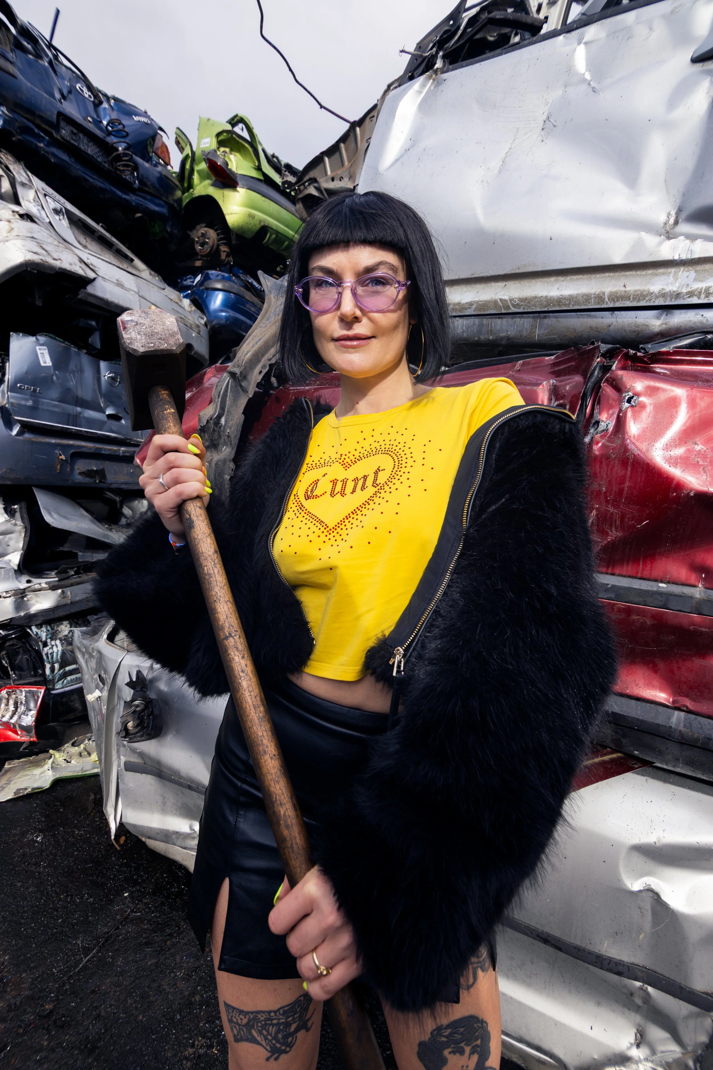 A woman with black hair, purple glasses, and a yellow t-shirt, holding a hammer, standing in front of a pile of wrecked cars.
