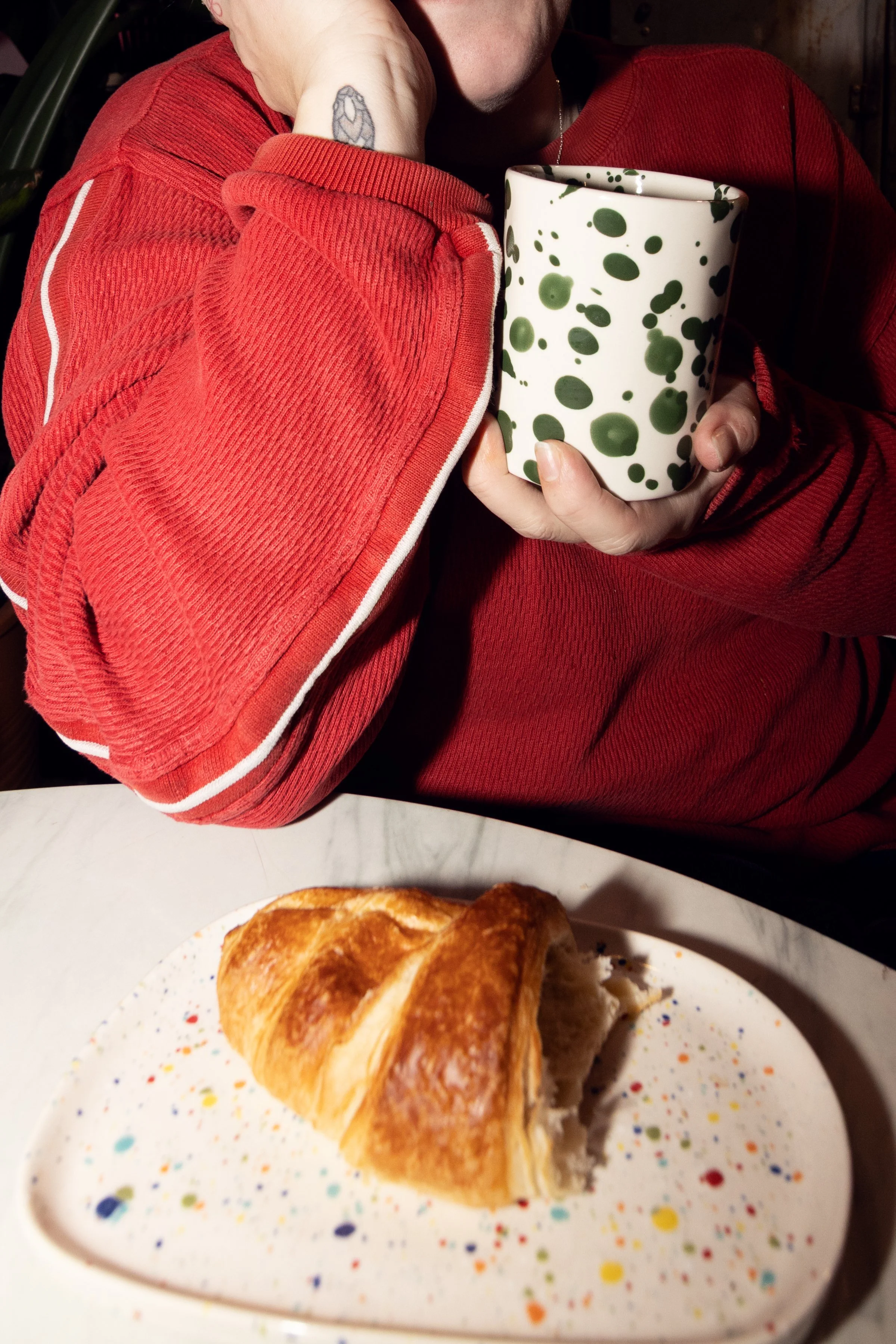 Person wearing a red sweatshirt holding a speckled green and white mug, with a croissant on a plate in front of them on a marble table.
