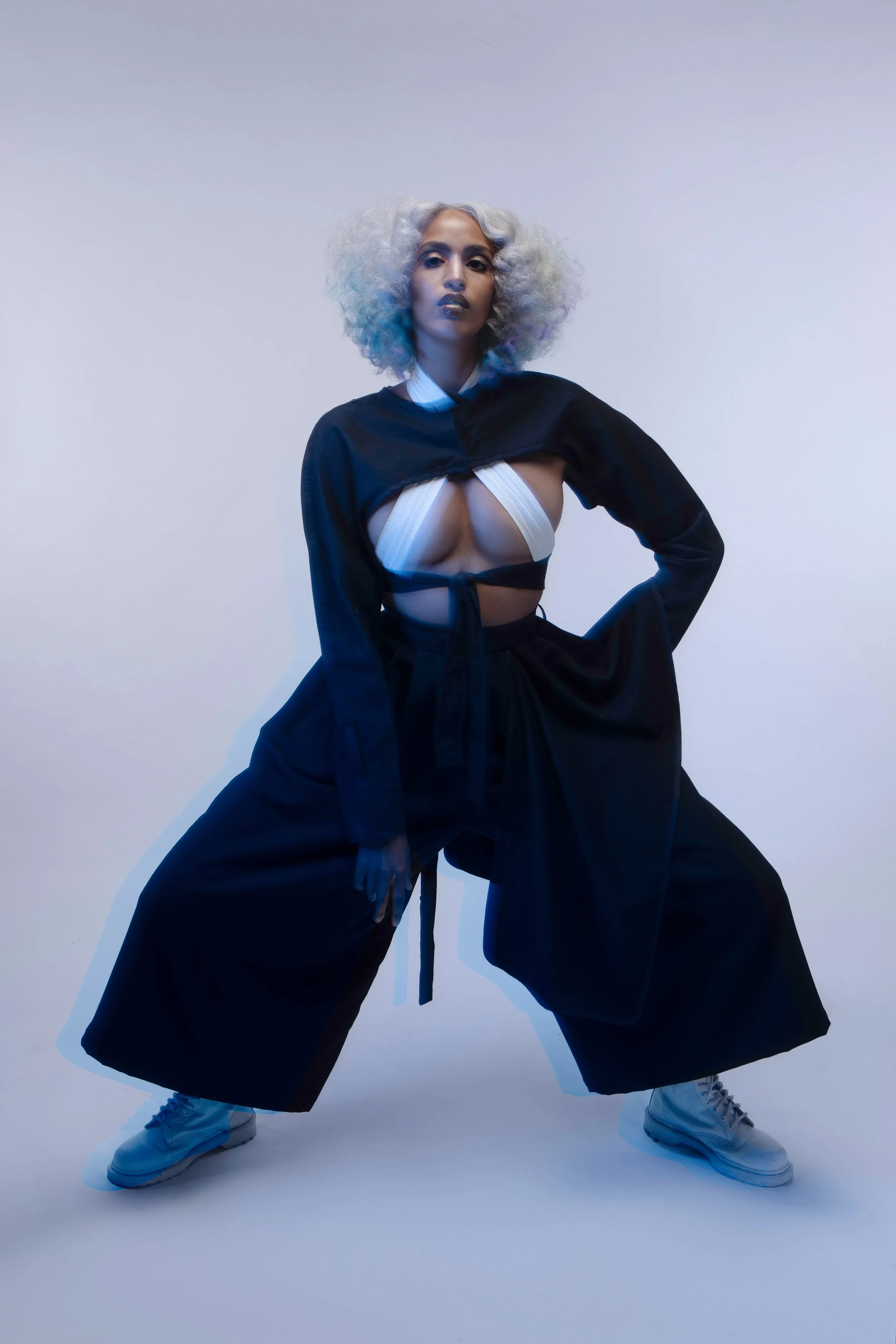 Fashion model with curly, platinum blonde hair posing in a studio against a plain background, wearing an avant-garde outfit with wide-legged pants, a black and white layered top, and sneakers.