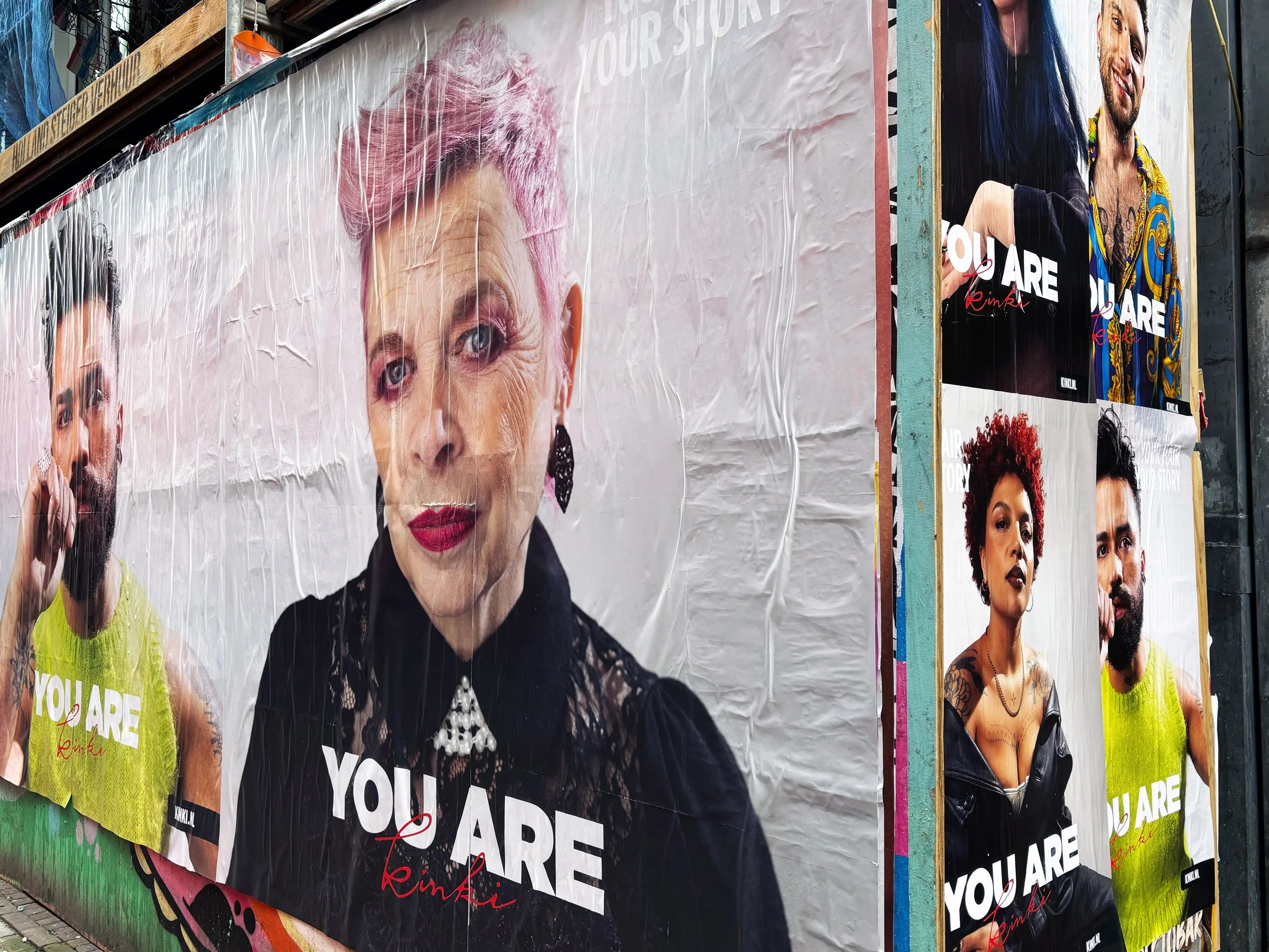 A large outdoor billboard featuring a woman with pink hair and red lipstick, wearing a black lace top and earrings, with the text "YOU ARE" and a signature "Rinki" in red. Surrounding posters feature diverse people with various hairstyles and fashion