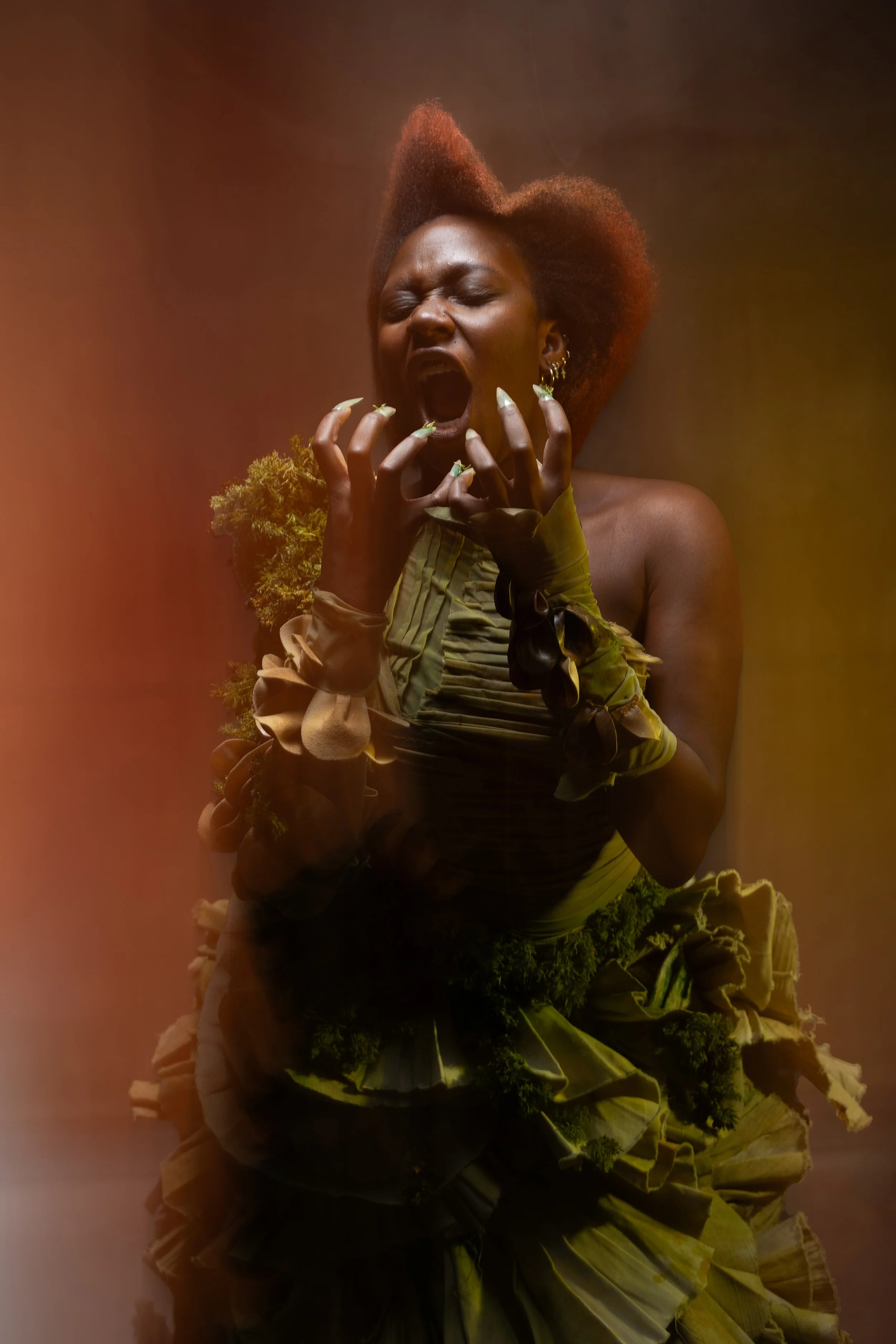 A woman with natural hair, dressed in a green outfit with ruffles and floral details, screams with her eyes closed and hands near her face, against a warm, gradient background.