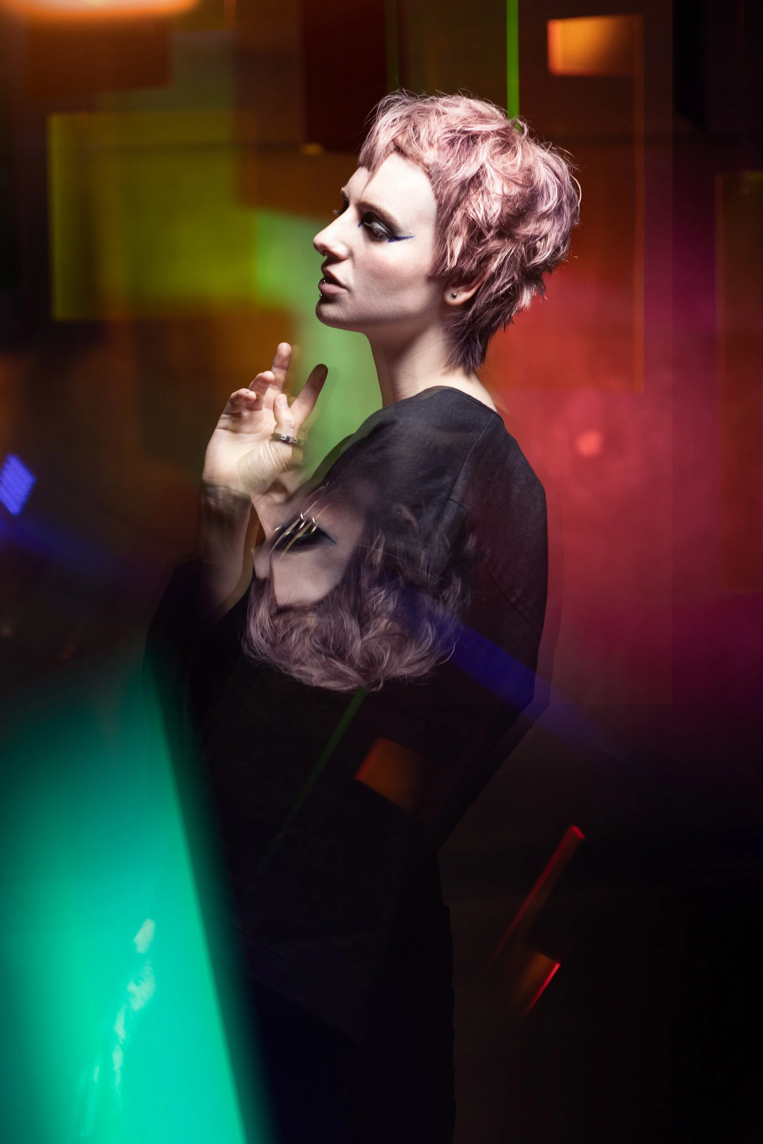 A woman with short, pink hair and dark makeup stands in front of colorful, abstract lighting. She is wearing a black top and has a contemplative expression.