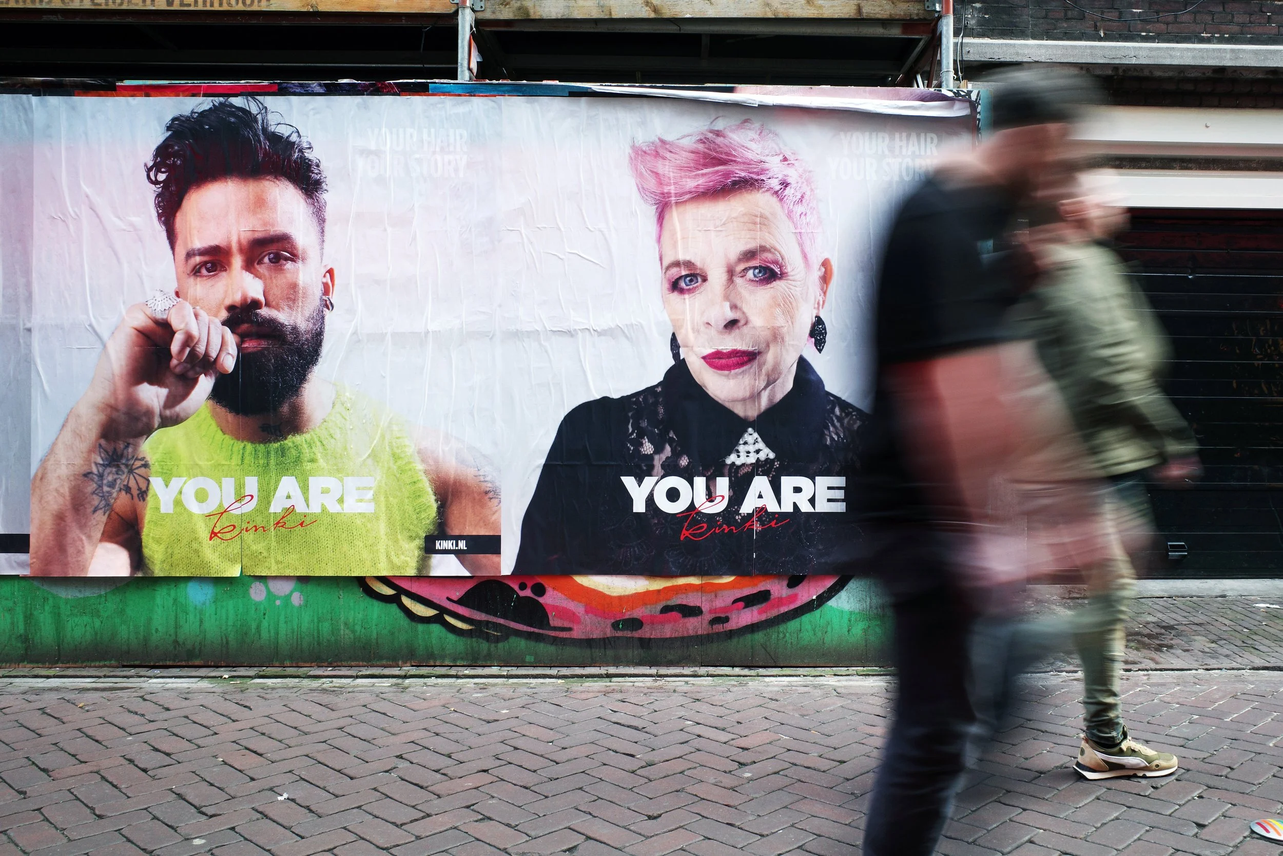 A city street with a large billboard featuring two portraits, one of a bearded man with dark hair and tattoos, wearing a green tank top, and the other of an older woman with pink hair and earrings. The billboard has the text "YOU ARE" above each port