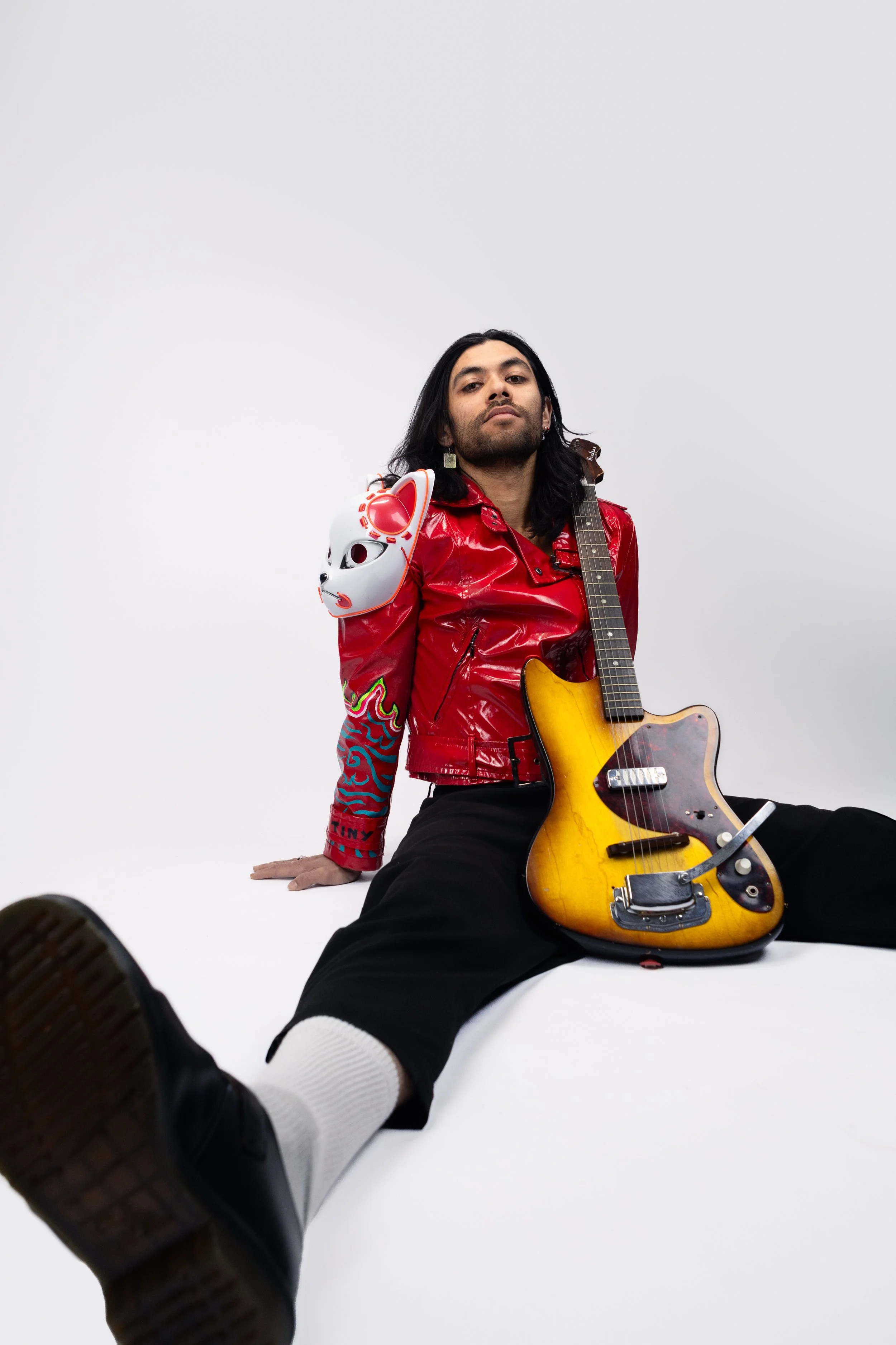 A person with long dark hair wearing a red shiny jacket, black pants, white socks, and black shoes, sitting on the floor against a plain white background with a yellow electric guitar on their lap and a white mask hanging from their shoulder.