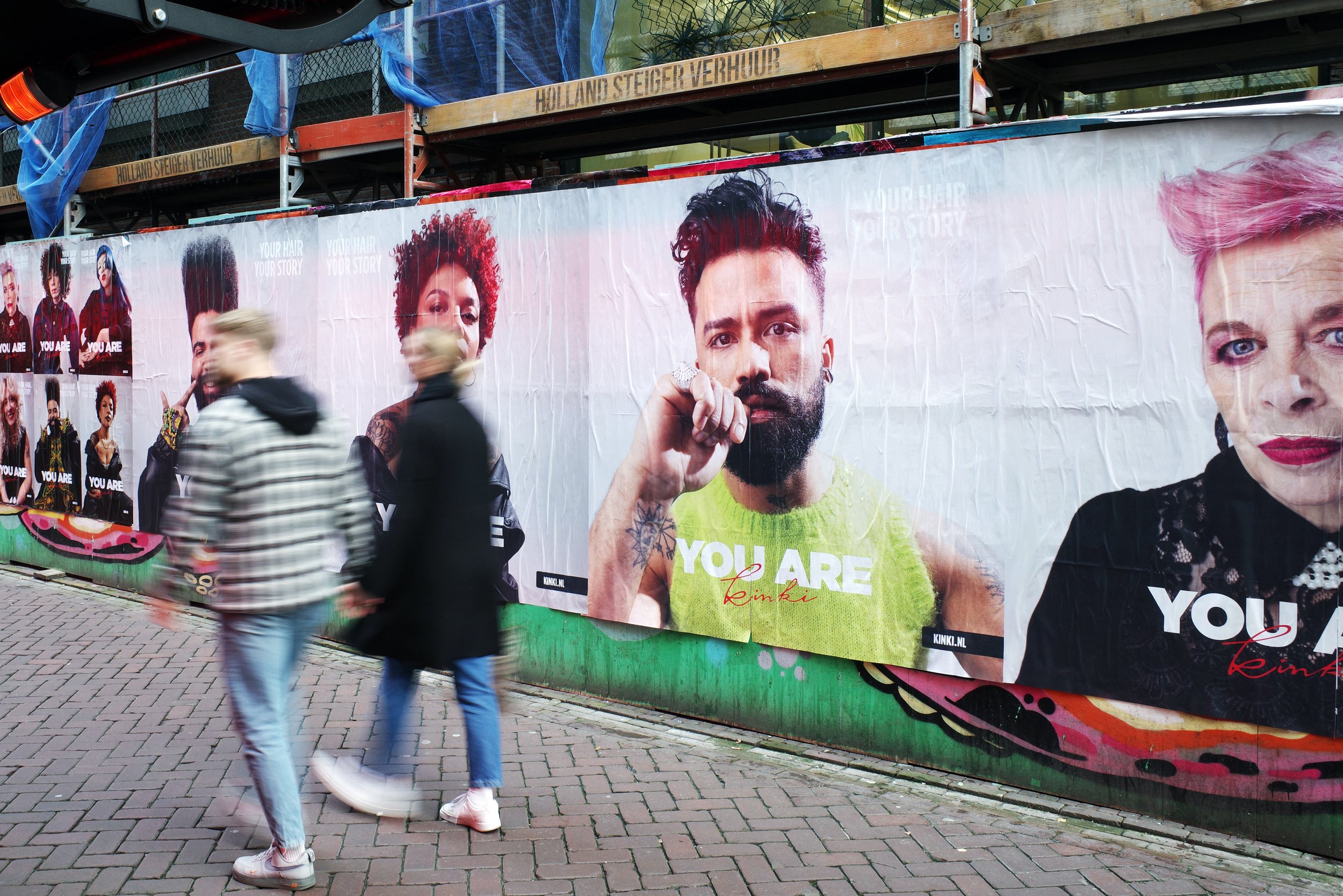 Street scene with blurred pedestrians walking past a large advertisement billboard featuring diverse faces and the phrase "YOU ARE" in bold letters.