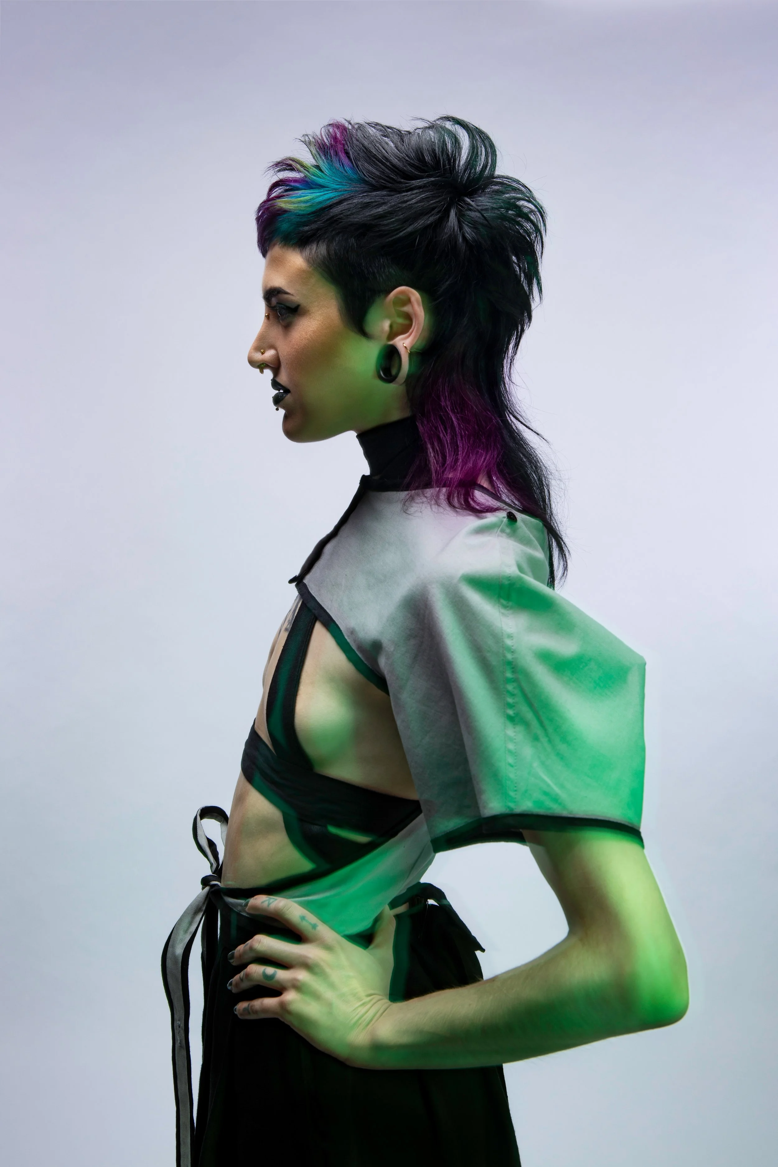 A woman in profile with a mohawk hairstyle dyed with blue, green, purple, and pink streaks, wearing black lipstick, earrings, and a futuristic outfit with sheer fabric and black accents.