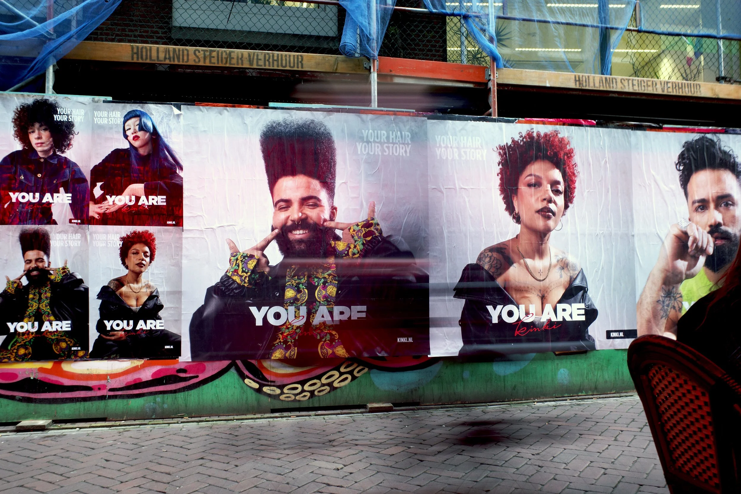 A large outdoor advertisement featuring diverse models with colorful hairstyles and tattoos, with the phrase 'YOU ARE' prominently displayed in white text.