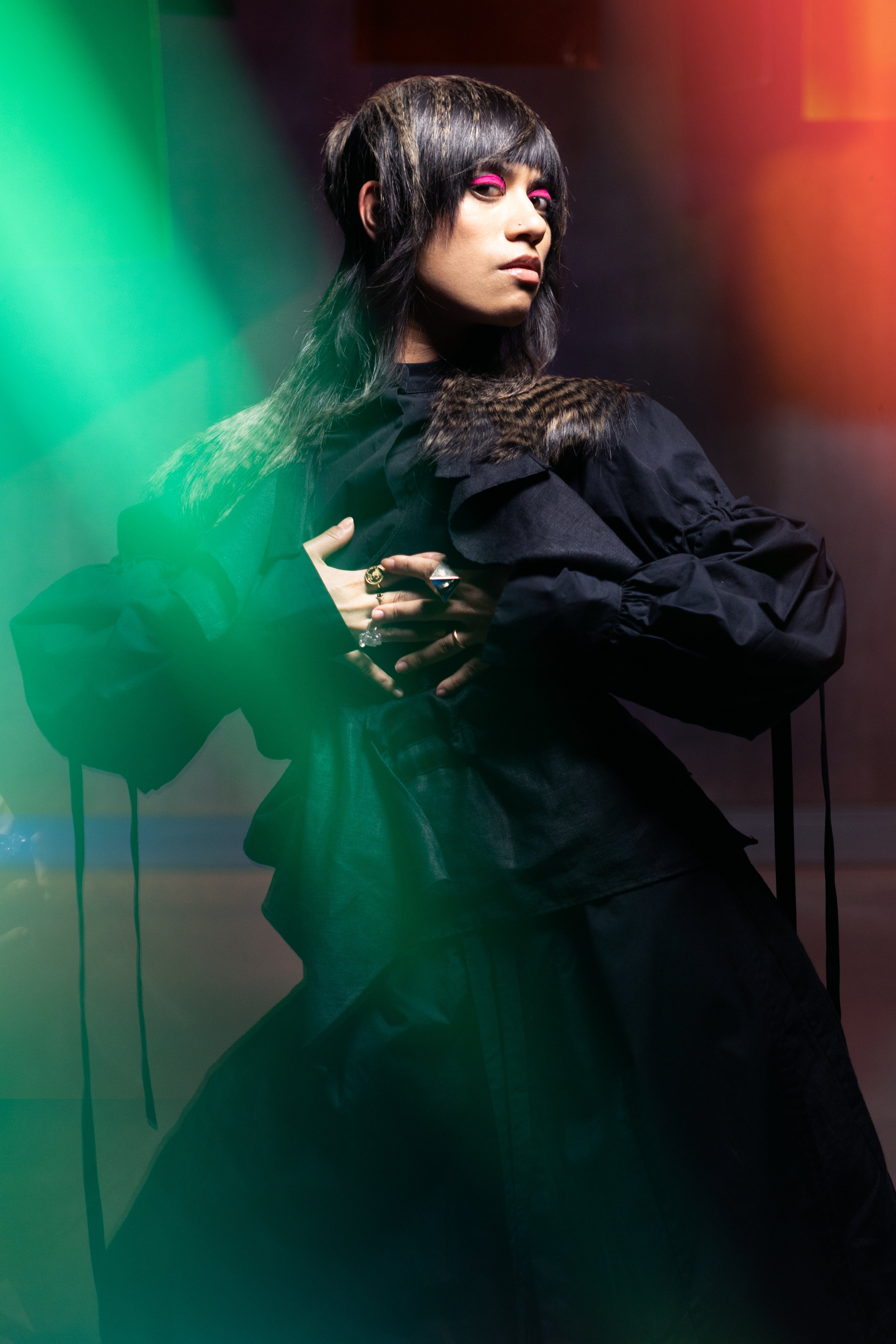 A woman with dark hair, pink eye makeup, and layered clothing poses against a colorful, gradient background that includes green, red, and yellow hues.