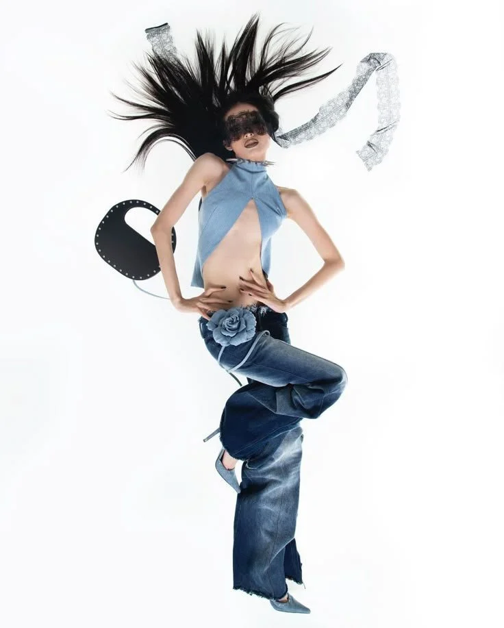 A person with long black hair, wearing a blindfold, a blue top with cutouts, jeans with a flower embellishment, and heels, posing dynamically against a plain white background.