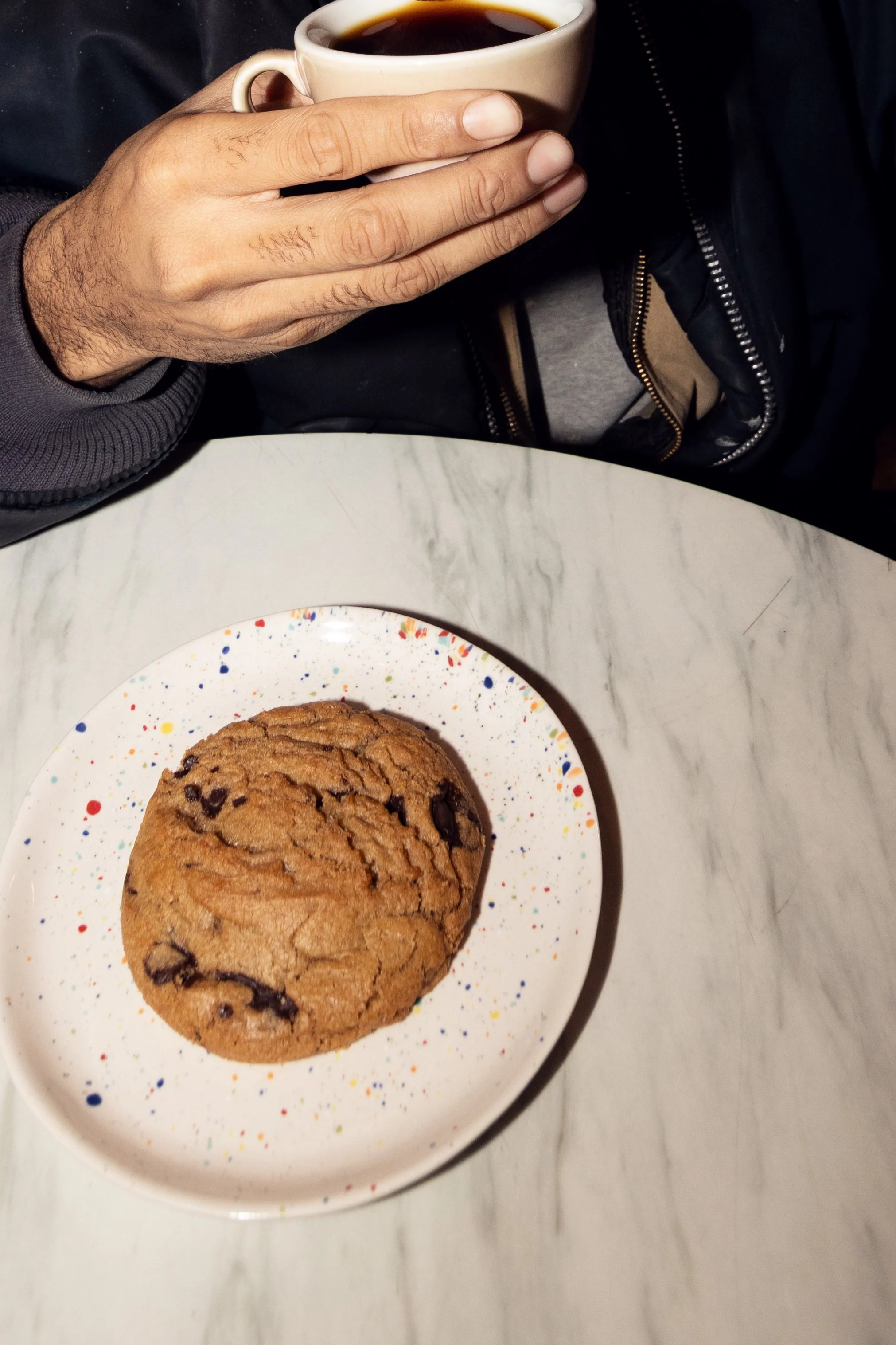 Person holding a cup of black coffee above a table with a chocolate chip cookie on a colorful splattered plate.