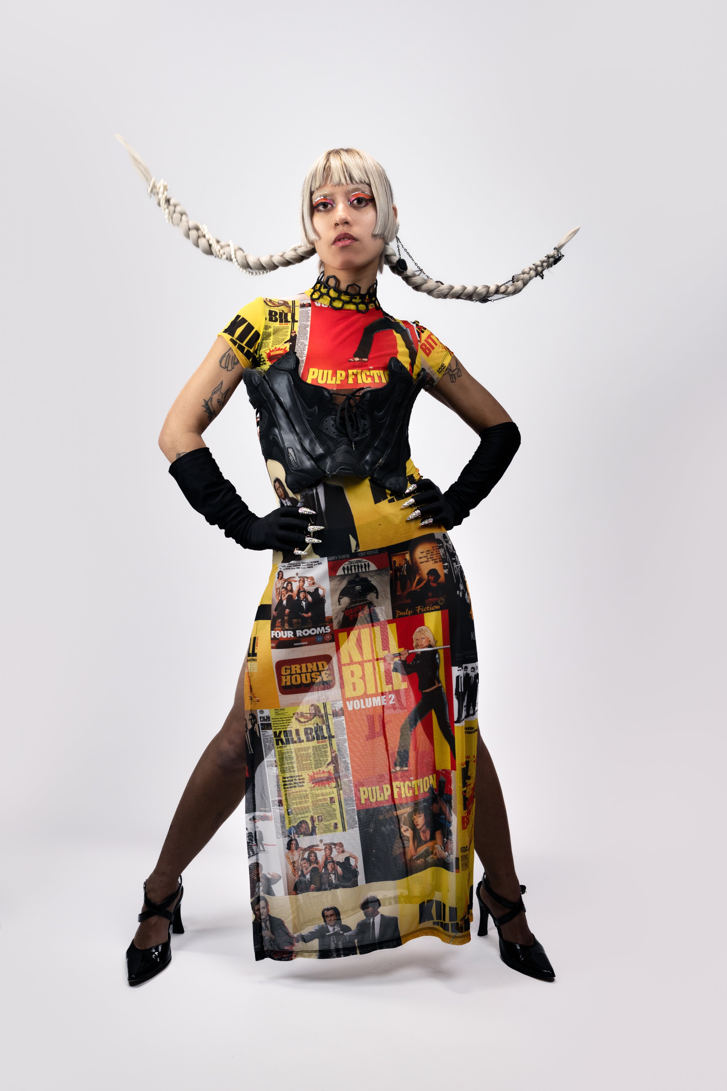 A woman with platinum blonde hair styled in long braided pigtails with pointed tips, wearing a colorful graphic dress with 'Pulp Fiction' and 'Kill Bill' themes, black gloves, a black bra, and high heels, standing with hands on hips against a plain w