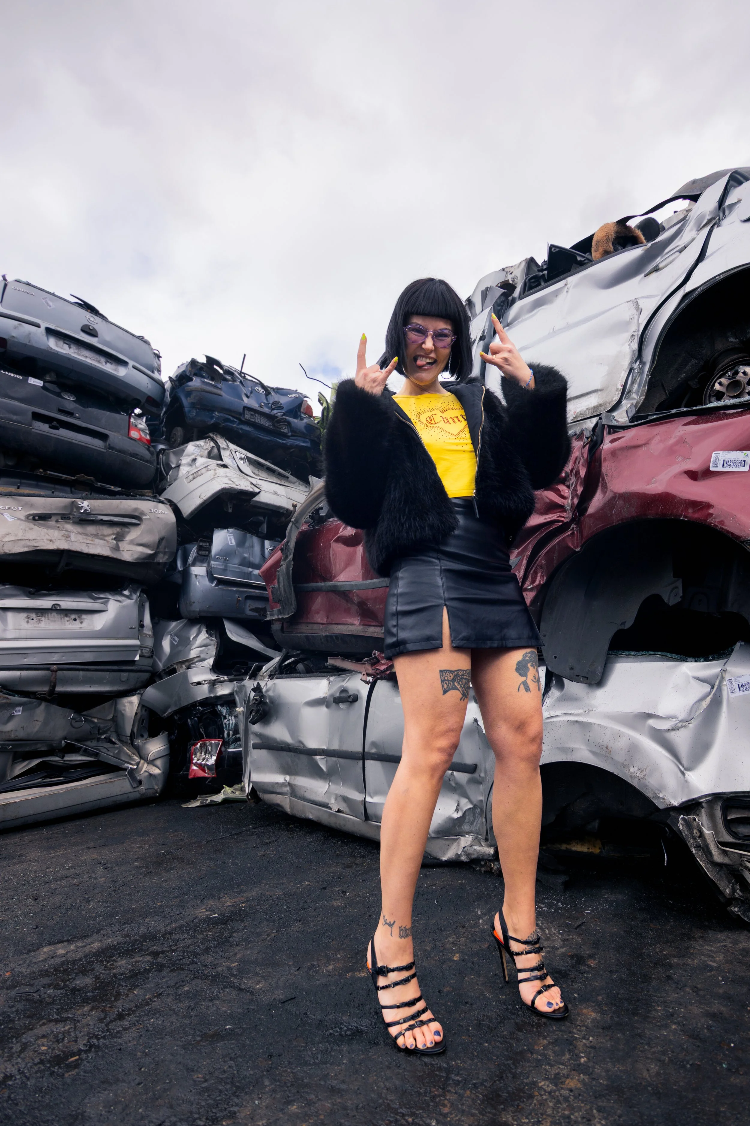 A woman with tattoos and black hair, wearing a yellow shirt, black fur jacket, and black leather skirt, standing in front of a pile of wrecked cars, making a rock and roll hand gesture.
